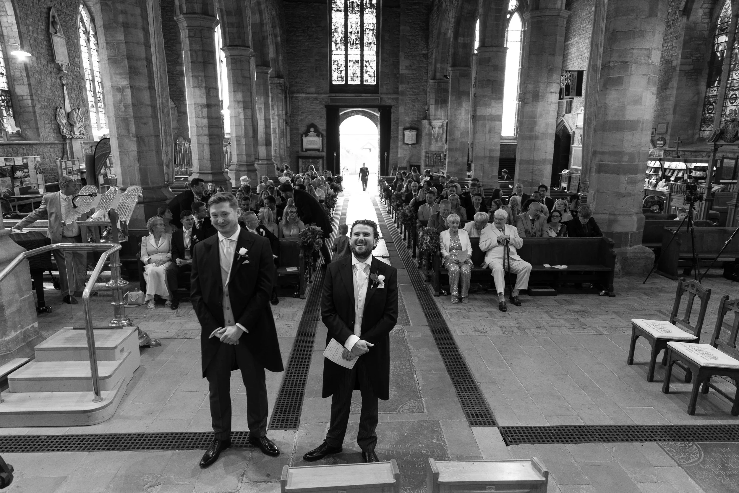 A wedding ceremony inside a large stone church with high stained glass windows. The audience is seated, and two men in tuxedos stand at the altar, smiling, with one holding a paper.
