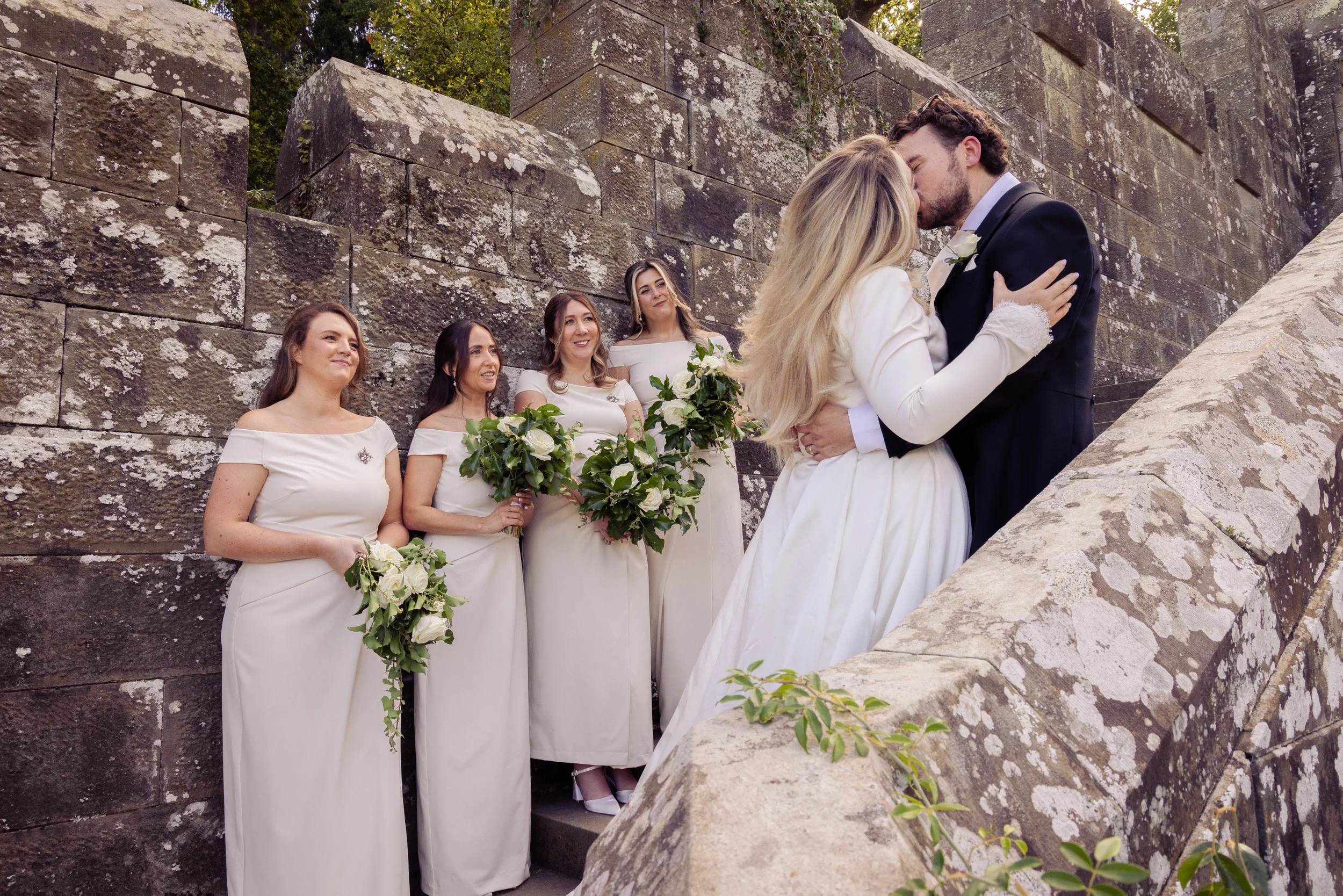 A wedding scene with a bride and groom kissing, surrounded by bridesmaids holding bouquets, against an old stone wall.