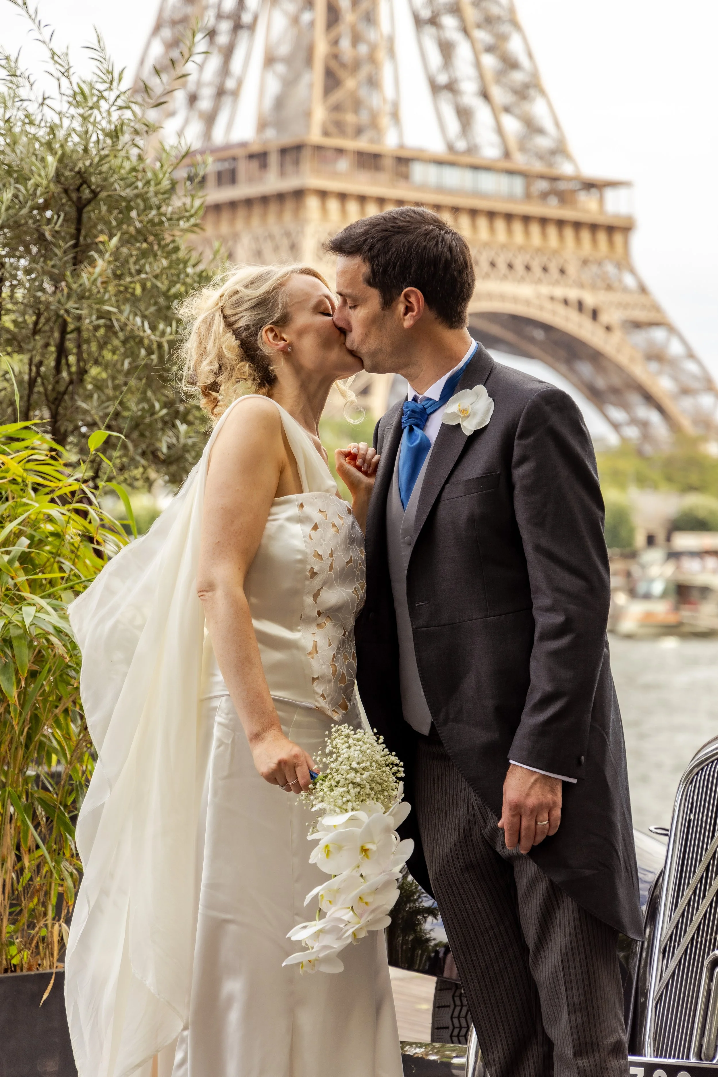 A bride and groom kissing in front of the Eiffel Tower, with greenery on one side and a boat with a wire chair in the foreground.