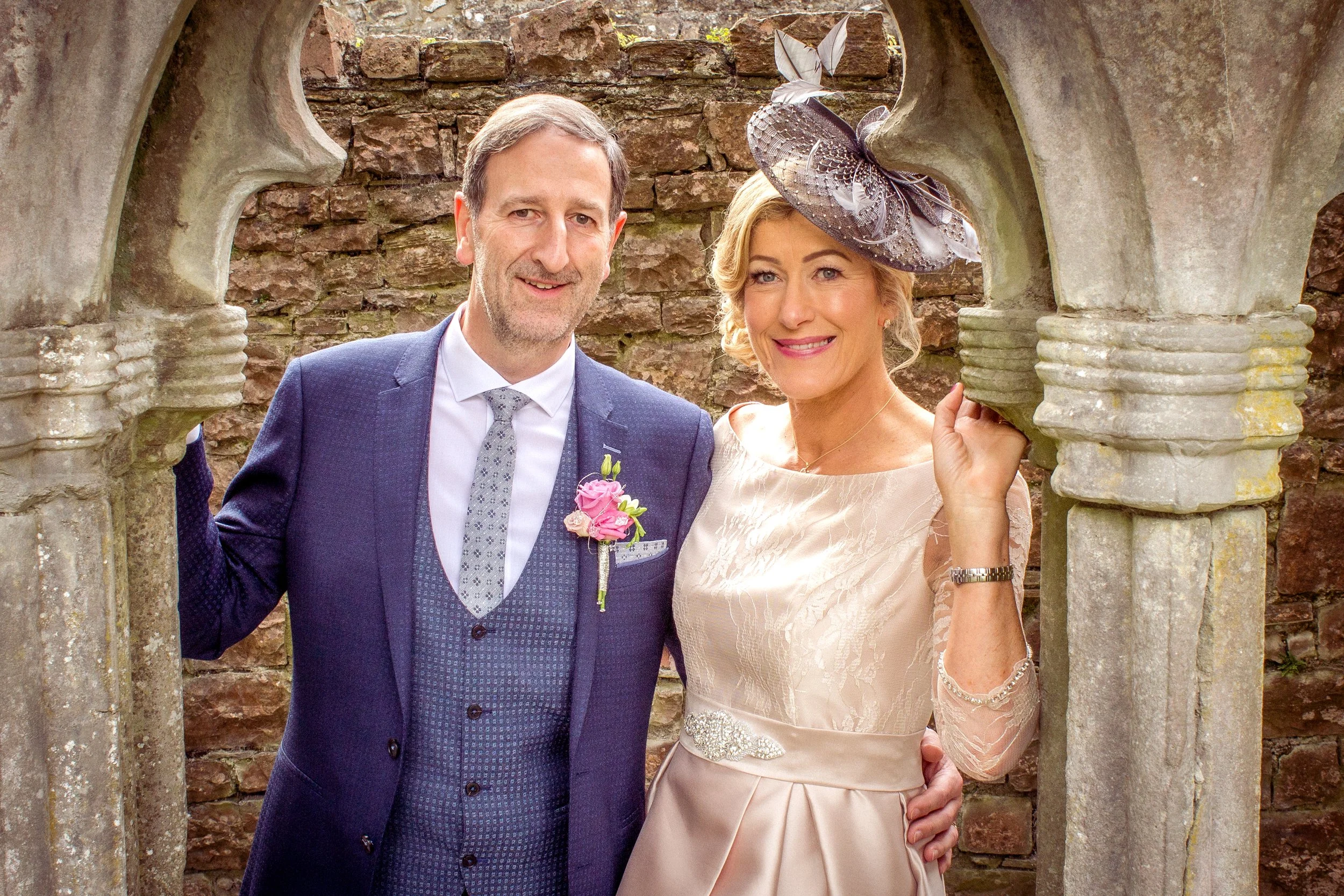 A happily married couple dressed in formal attire posing in a stone archway at an outdoor wedding venue.