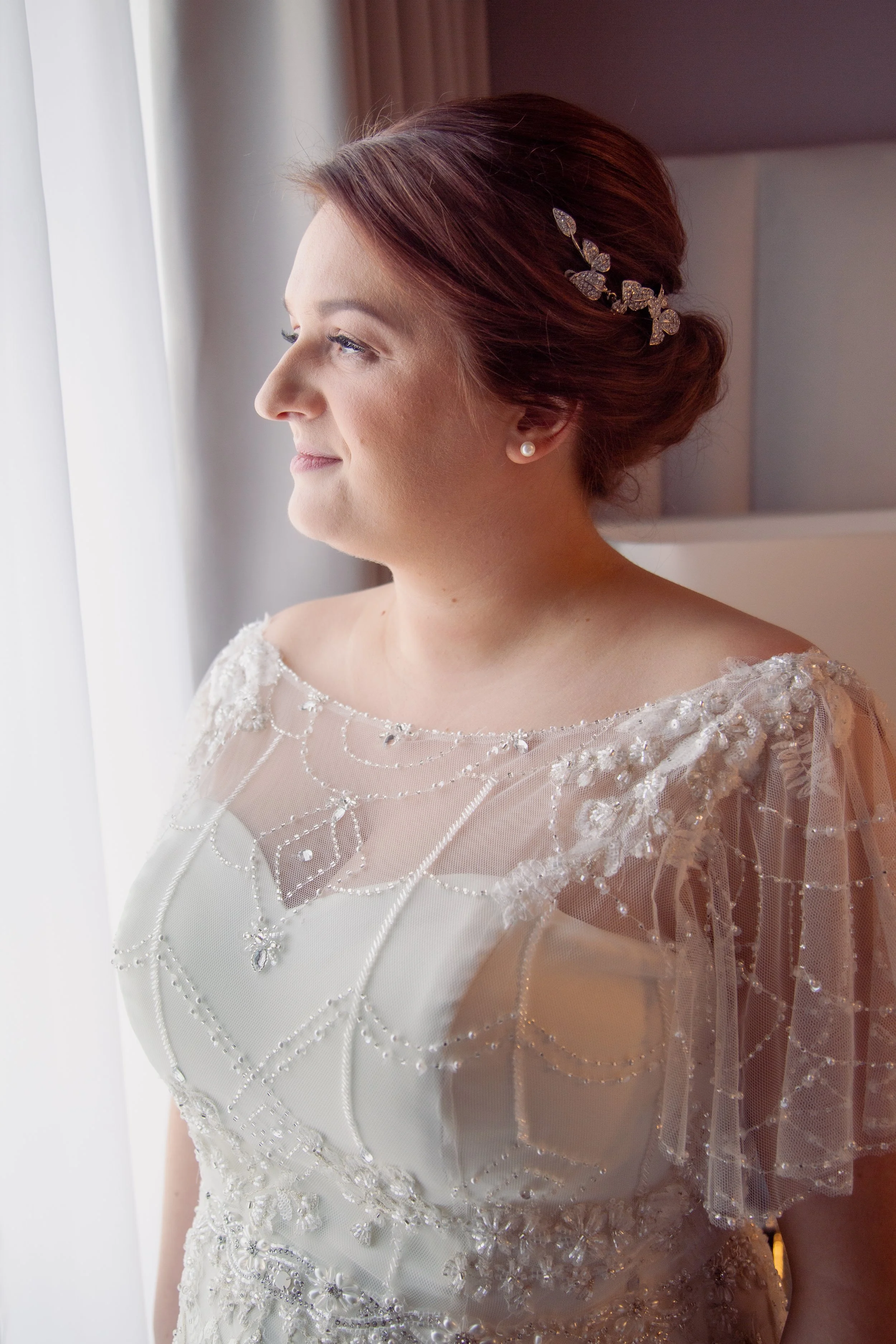 Close-up of a bride with red hair styled in an updo, wearing a pearl earring and a jeweled hair accessory, looking out a window, dressed in an elegant wedding gown with bead and lace embellishments.