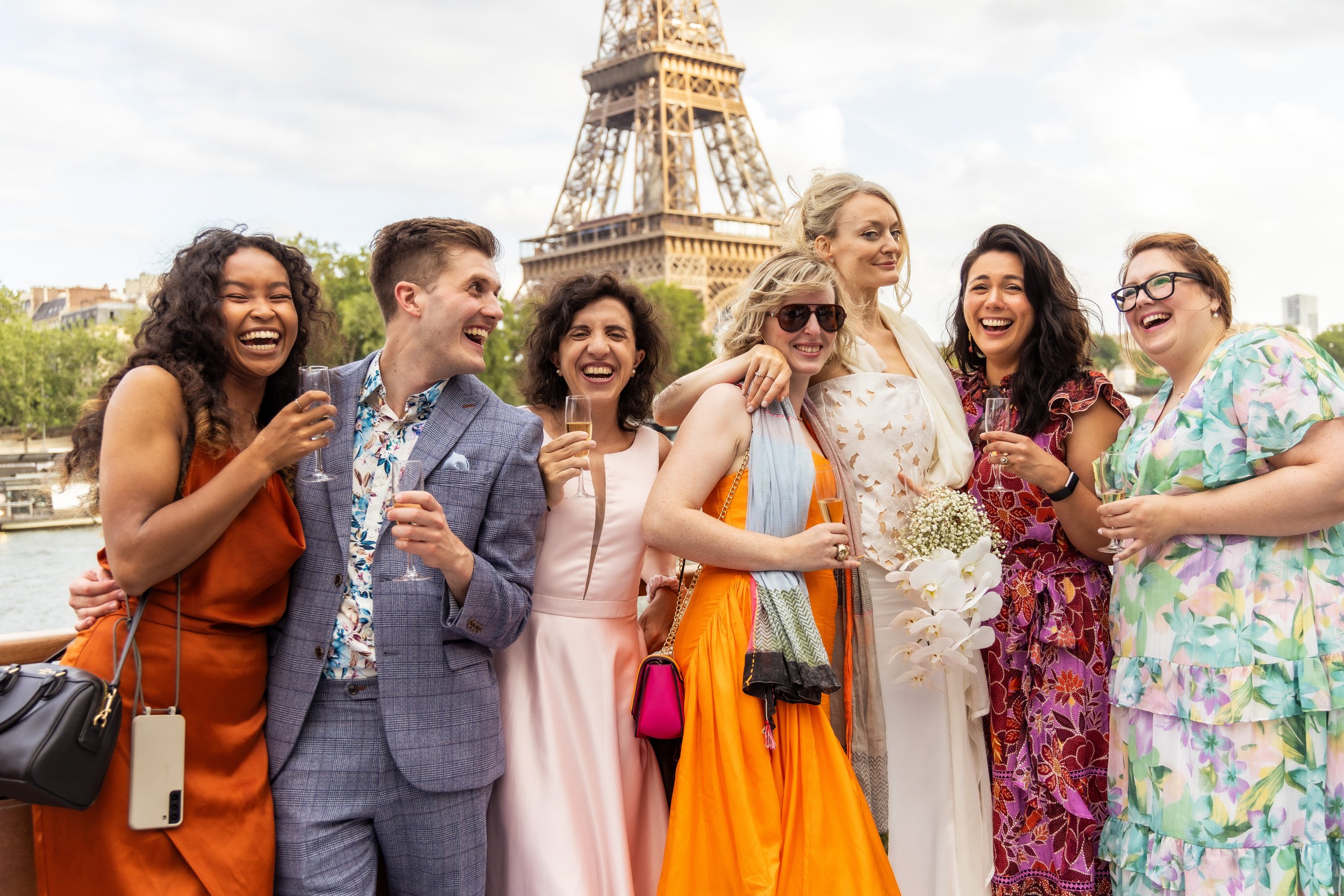 Group of seven women and one man smiling and enjoying drinks on a boat near the Eiffel Tower in Paris.