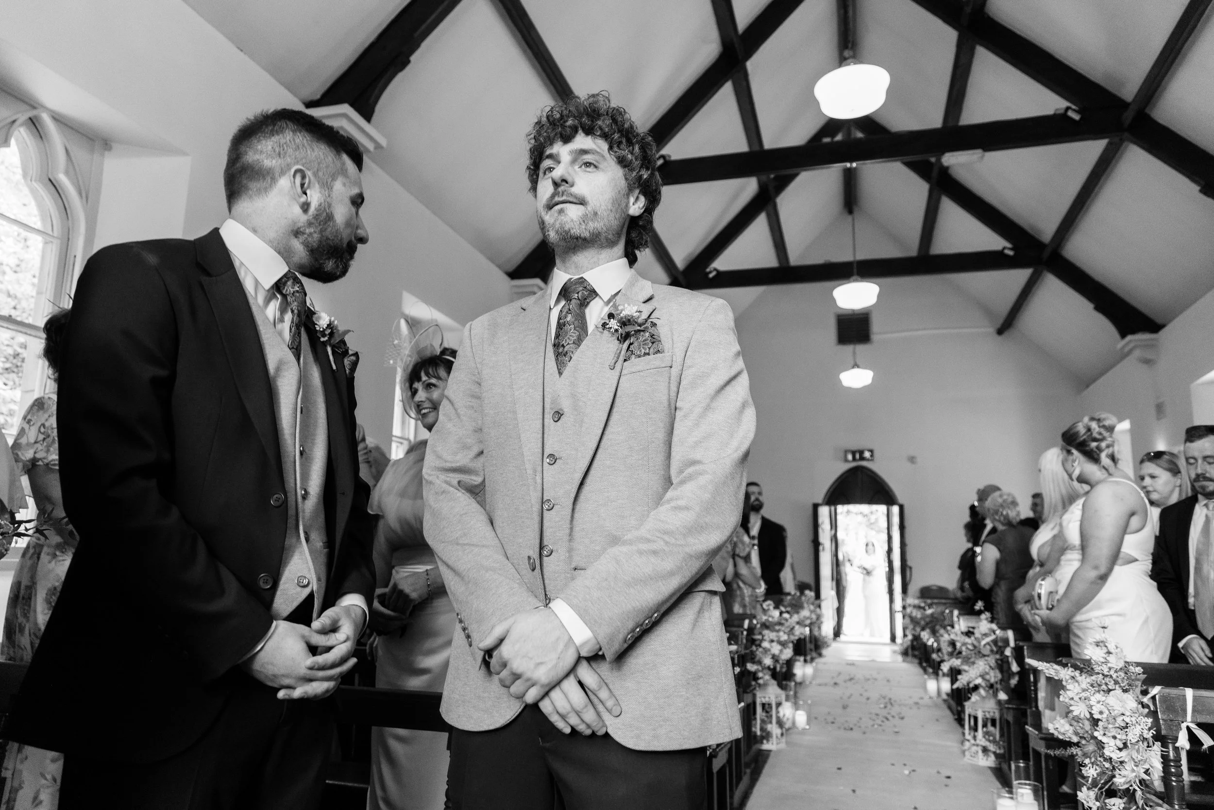 Black and white photo of a wedding ceremony with two men dressed in suits holding hands, standing in front of a church aisle with guests seated on both sides.