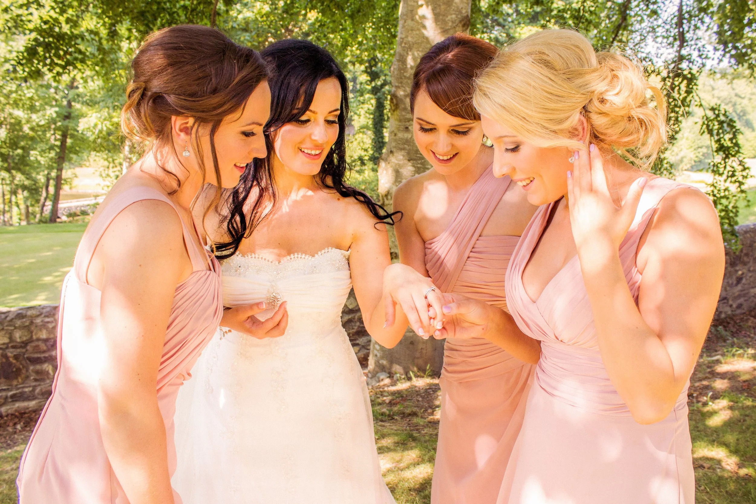 A bride and four bridesmaids gathered outdoors, smiling and looking at rings during a wedding celebration.