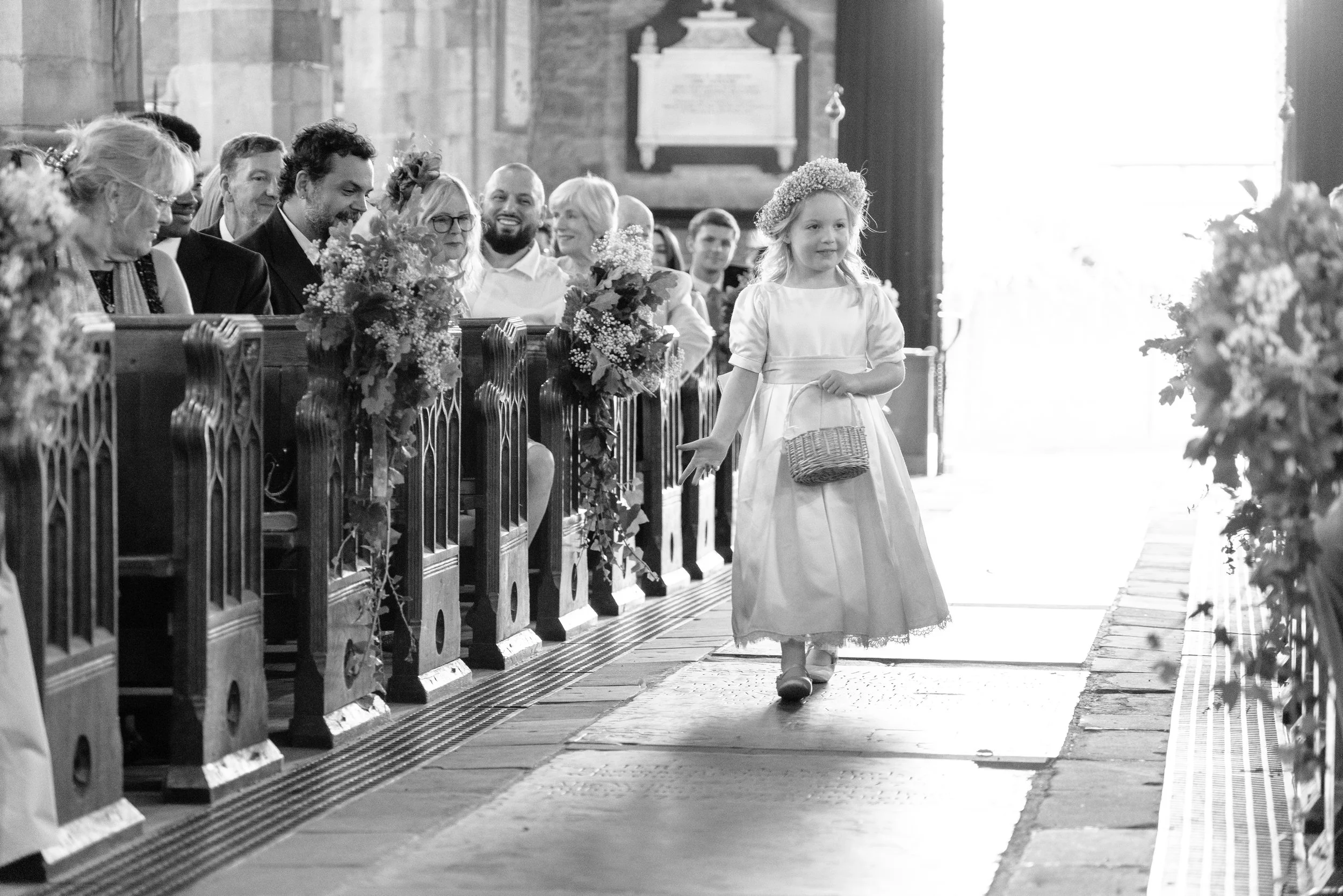 A young girl in a white dress and floral headband walking down the aisle in a church, carrying a small basket, with smiling adults seated on wooden pews decorated with flowers to her left.