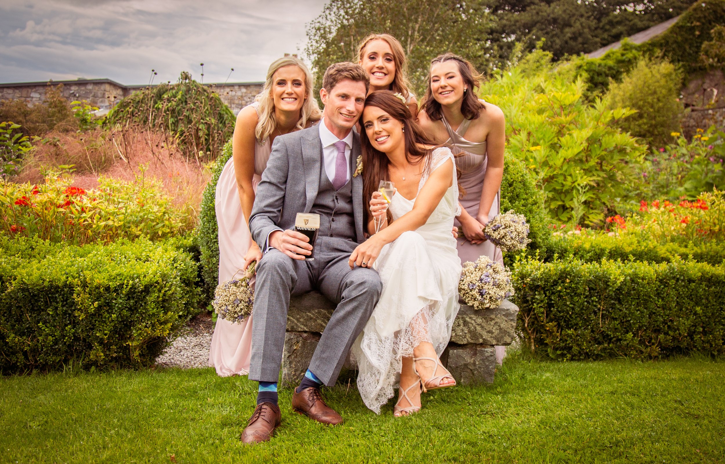 A group of six young adults, dressed in formal wedding attire, smiling and sitting on a rock in a garden. The bride is holding a glass of champagne, and a man next to her is holding a Guinness beer. They are surrounded by lush greenery and colorful flowers with an overcast sky in the background.