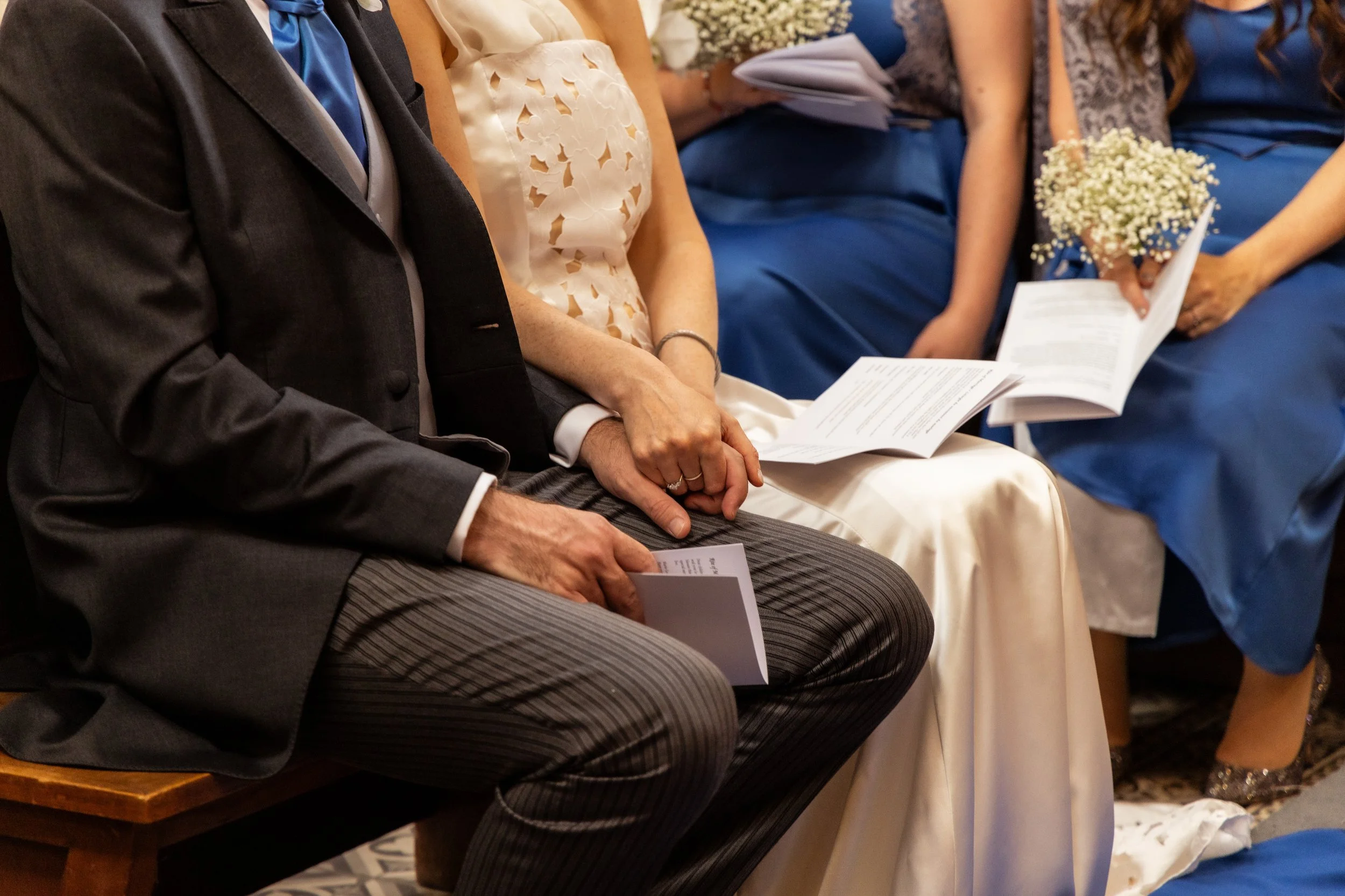 People sitting in the first row at a wedding, holding programs, with women in blue dresses, some holding bouquets of flowers, and one man in a suit.