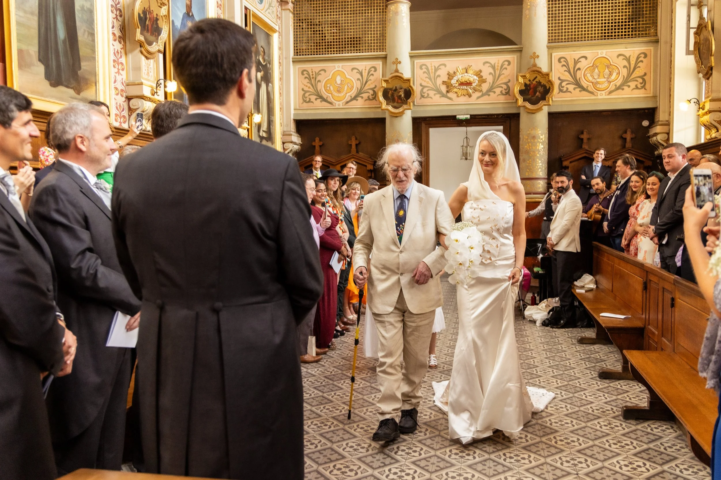 A bride in a white wedding dress walking down the aisle of a church escorted by an elderly man, possibly her grandfather, who is using a cane. The ceremony is attended by guests, some standing and taking photos, inside a decorated church with religio