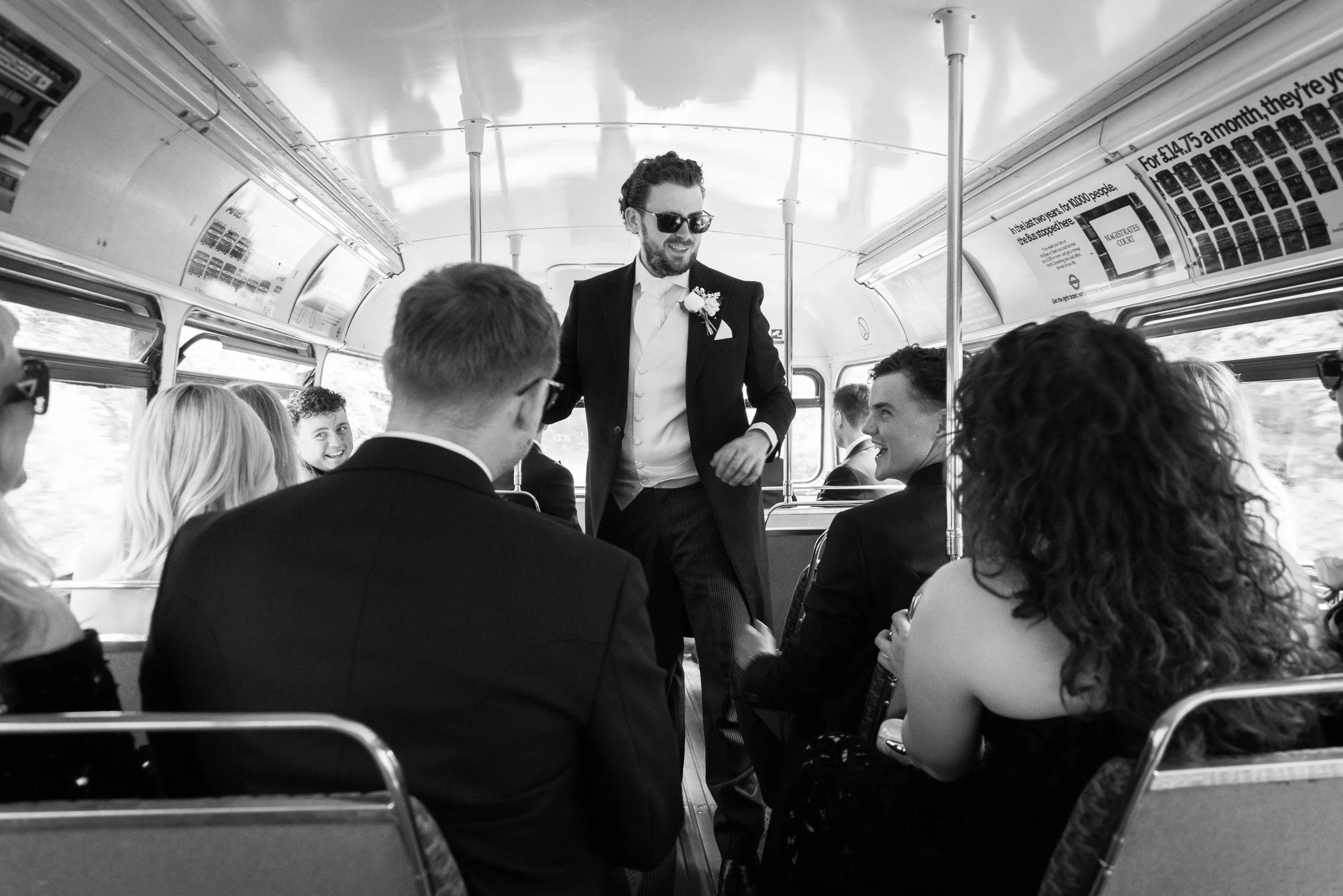 A black and white photo of a group of people on a bus, with a man in a tuxedo and sunglasses standing and smiling, possibly giving a speech, while the others seated among them are engaged and smiling.