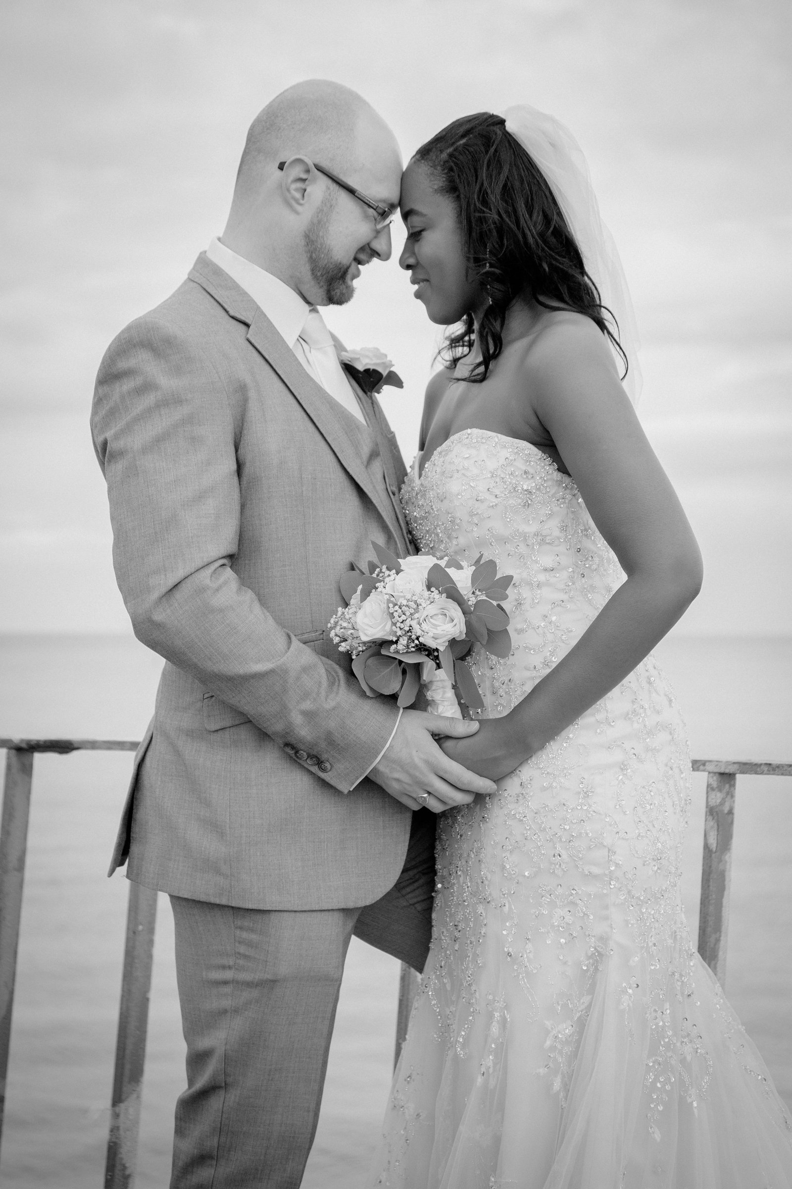A wedding couple, a man in a light-colored suit and a woman in a strapless wedding dress with intricate beading, holding hands with their foreheads touching, standing on a deck overlooking a body of water, in black and white.
