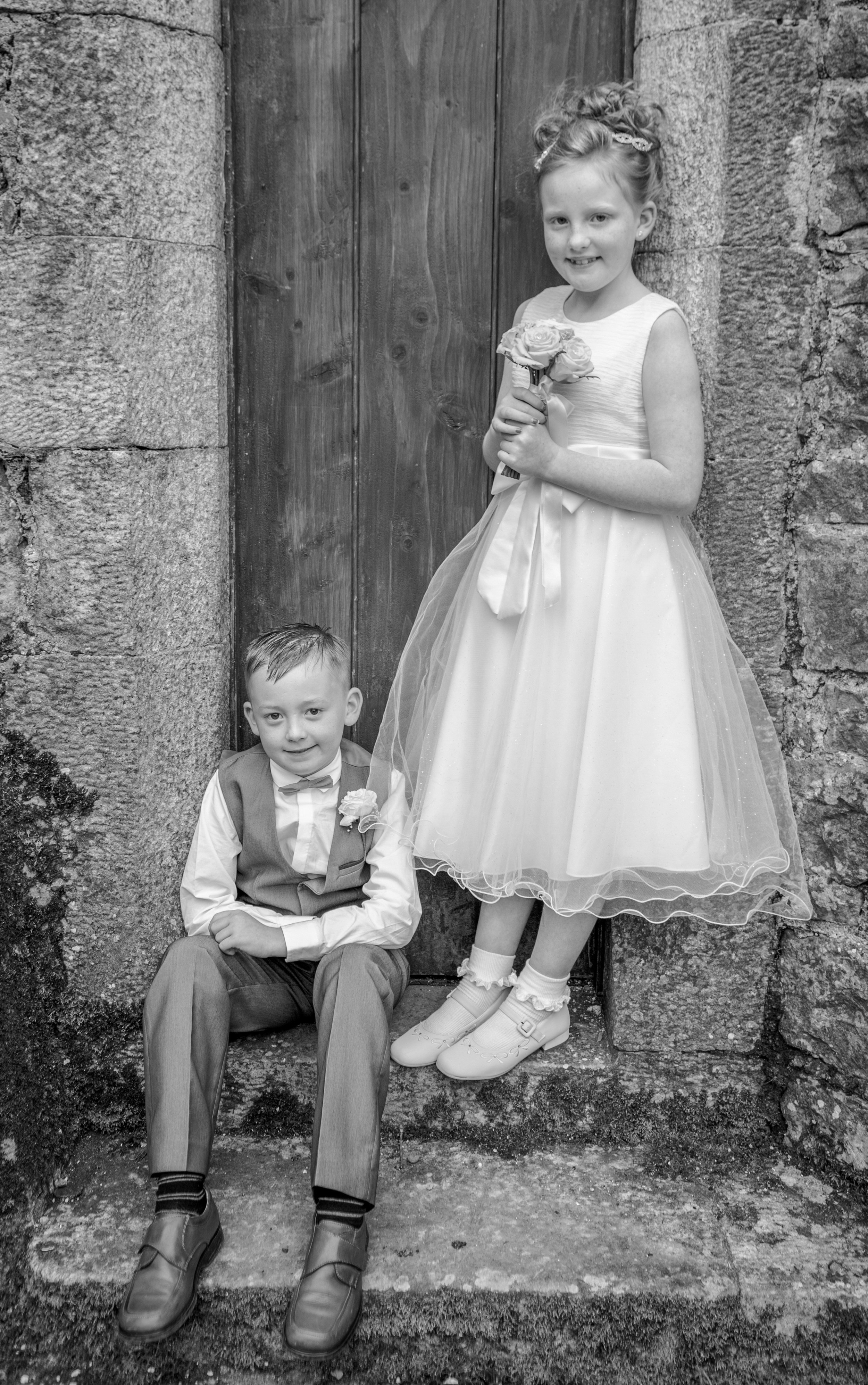 A young girl in a wedding dress holding a bouquet of flowers, standing next to a young boy seated on steps, both smiling, with a stone wall and wooden door in the background.