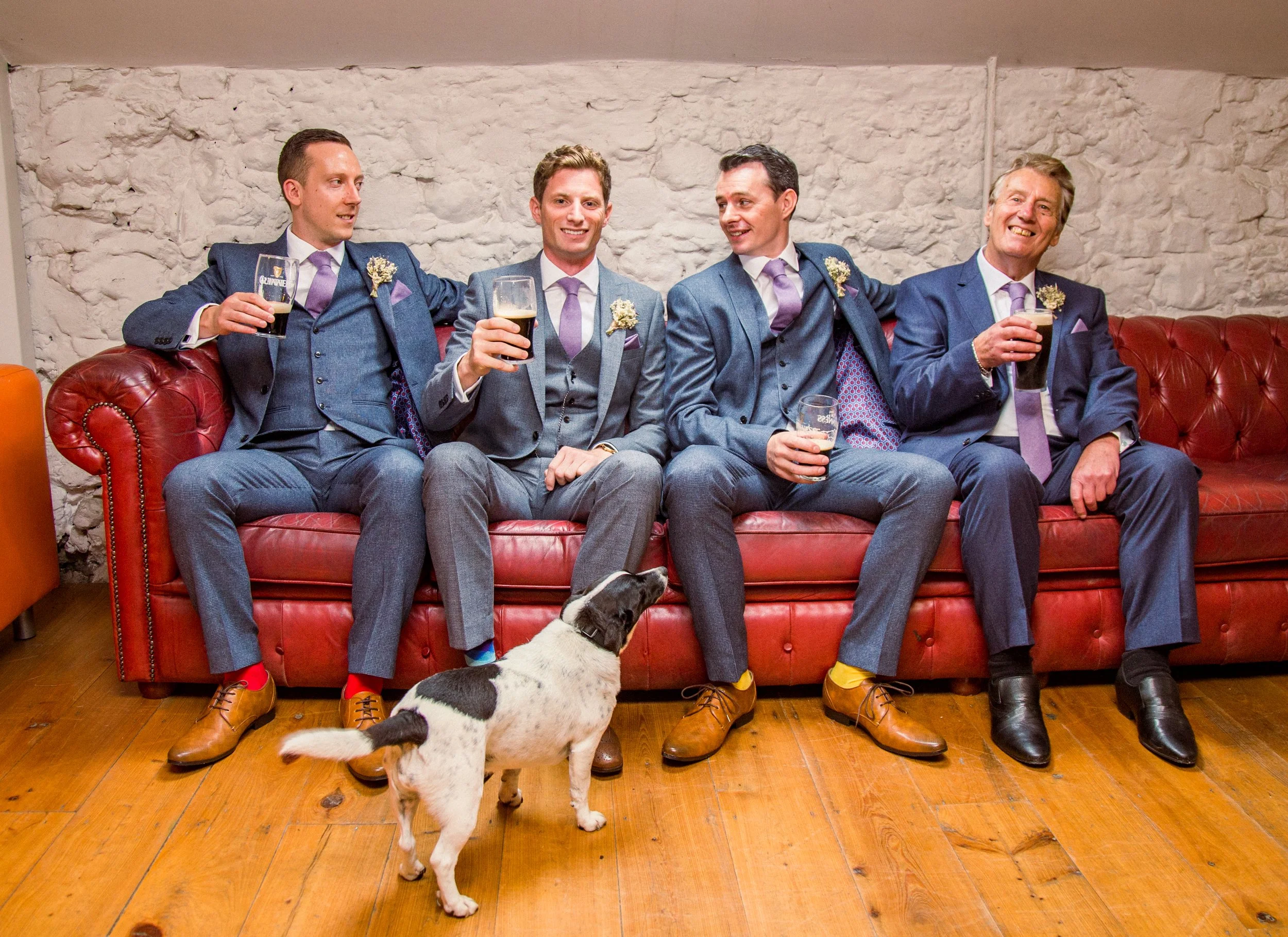Four men in suits and ties sitting on a red leather couch, holding glasses of dark beer, with two decorated with floral boutonnieres, and a black and white dog standing in front of them on a wooden floor in a room with white brick walls.
