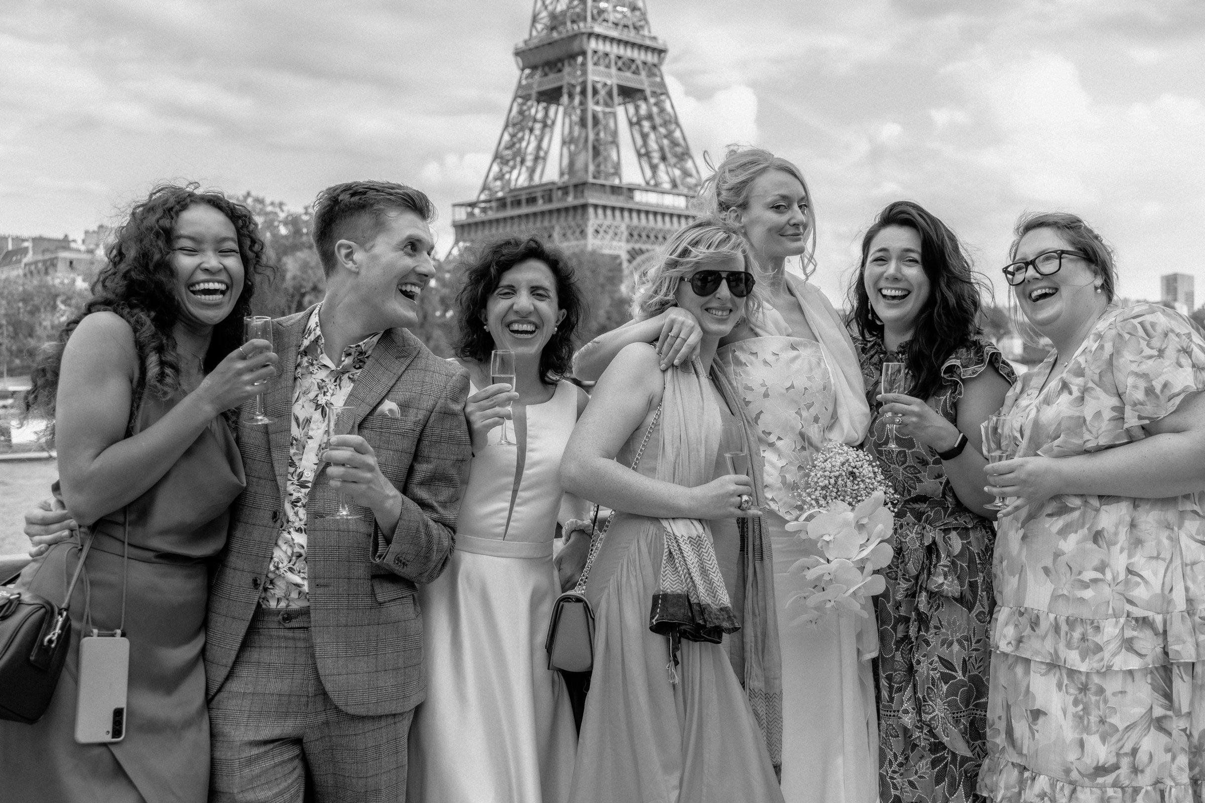 A group of seven diverse women smiling and laughing together outdoors, with the Eiffel Tower visible in the background, some holding wine glasses and one holding a bouquet of flowers.