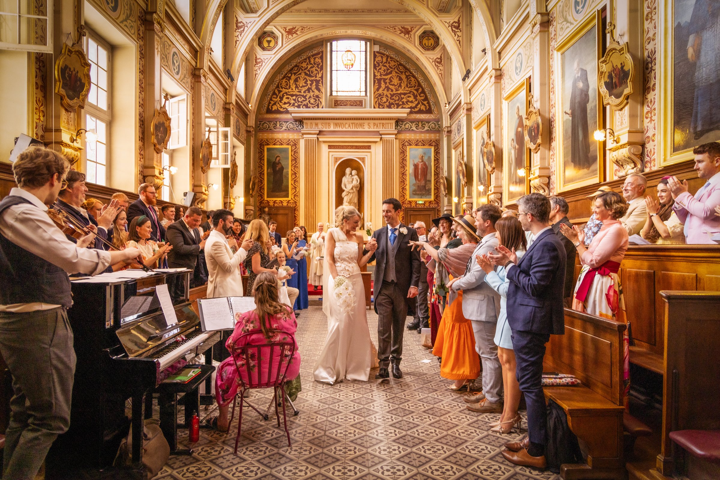 A wedding ceremony inside a decorated church with a bride and groom walking down the aisle surrounded by family and friends, with a pianist playing on the left.