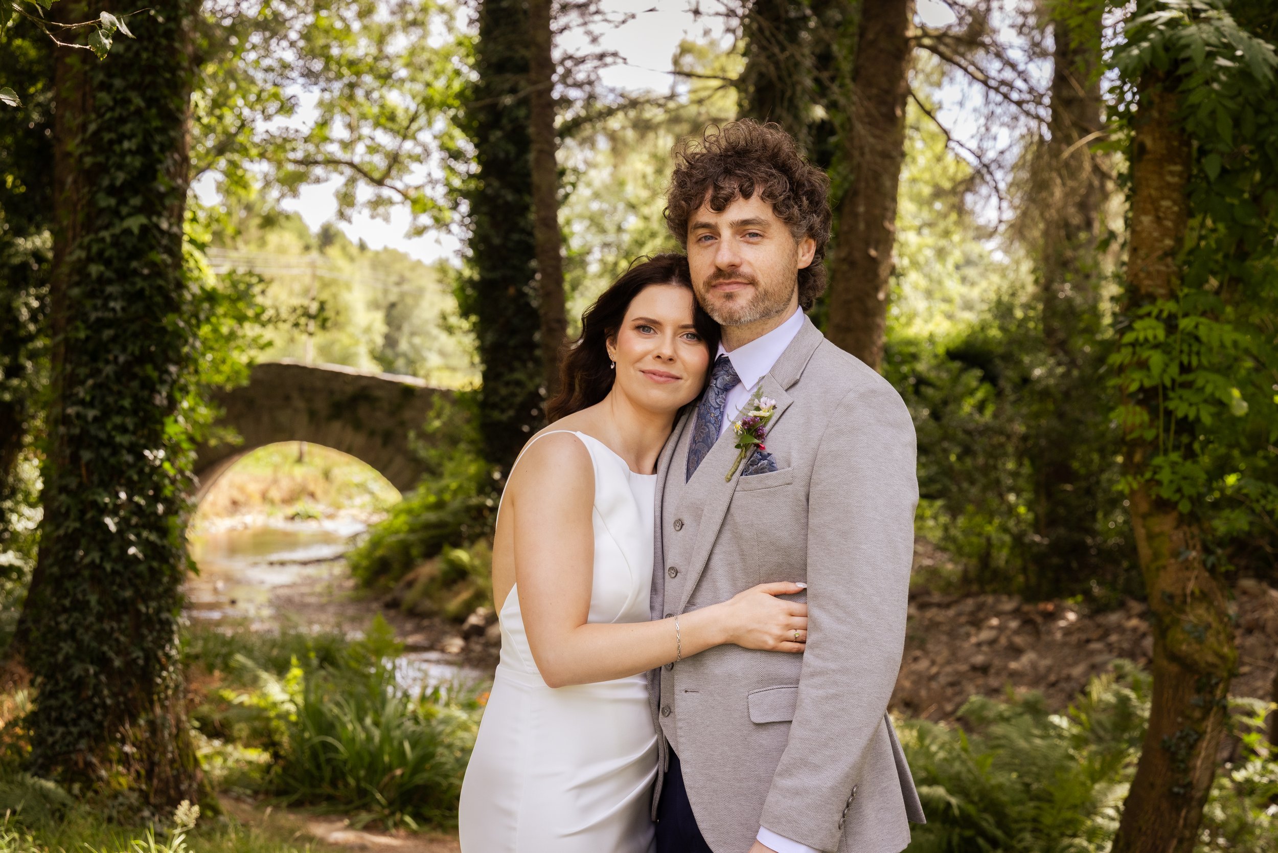 A man and woman dressed in wedding attire standing close together outdoors among trees and greenery, with the woman resting her head on the man's shoulder.