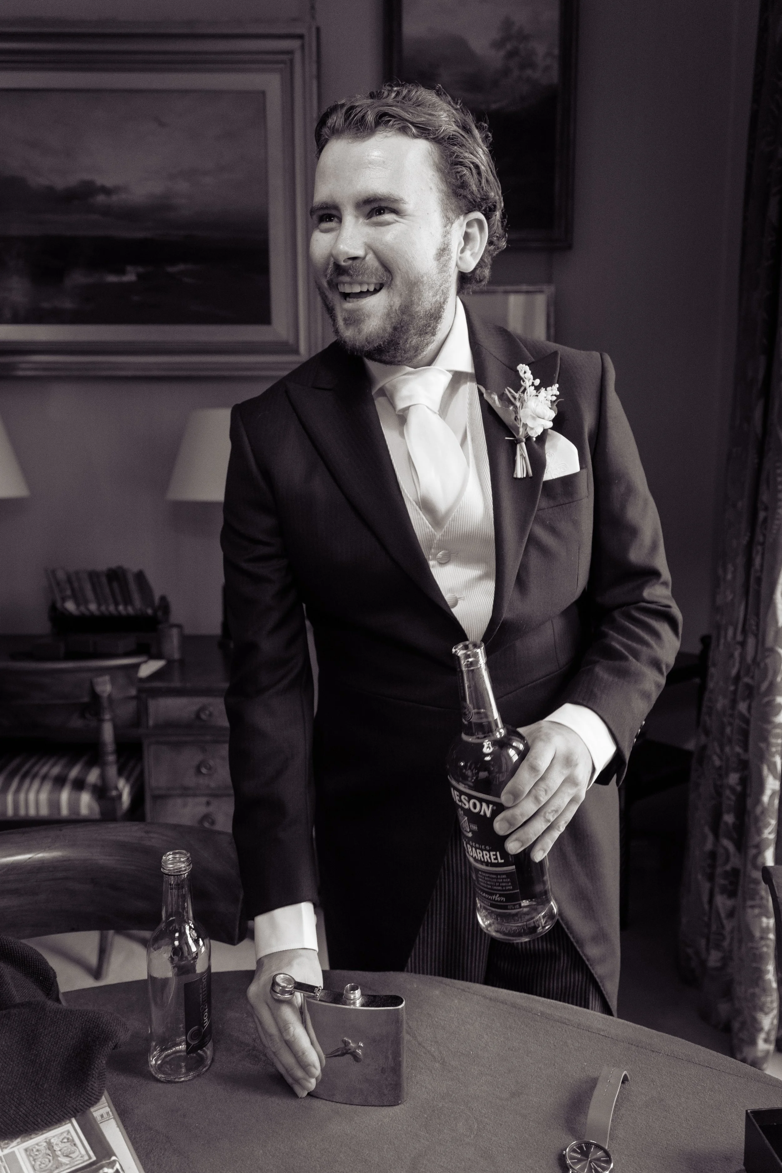 A man in a suit with a boutonniere is smiling and holding a liquor bottle while preparing a craft cocktail in a dimly lit room.