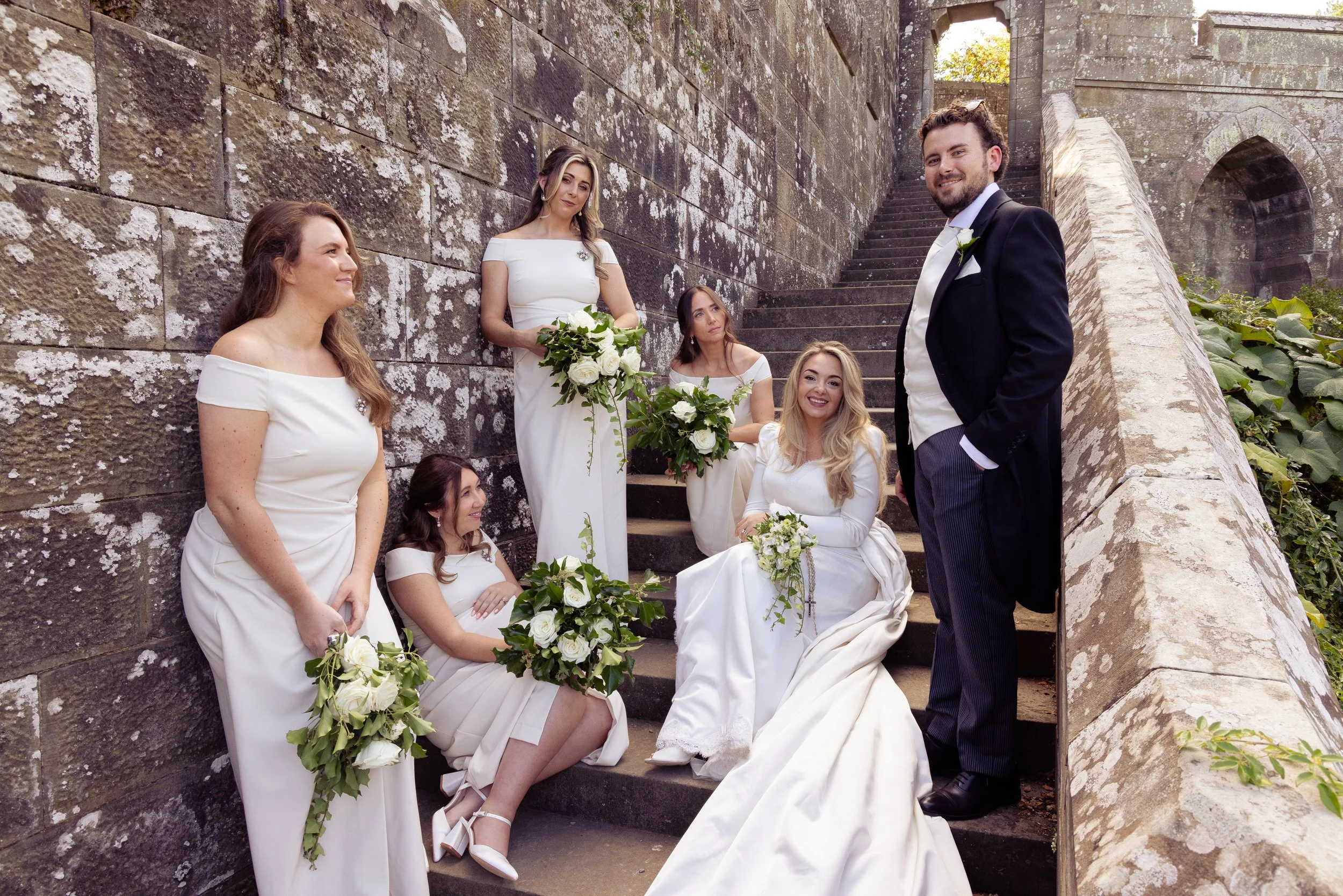 Group of women in white dresses sitting on stone steps with a man in a tuxedo standing nearby, outdoors near a stone wall, during a wedding celebration.