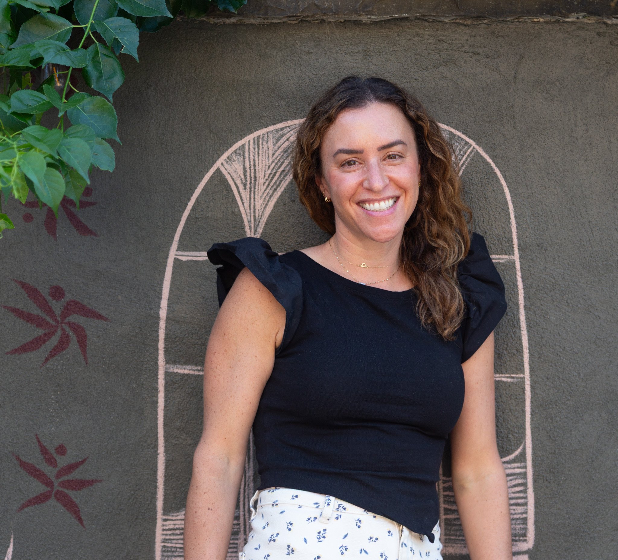 Dr. Sophia Brandstetter, LCSW, PsyD smiling, wearing a black top, with her head leaning back against a gray wall that has a pastel pink circular design and some green leaves in the top left corner.