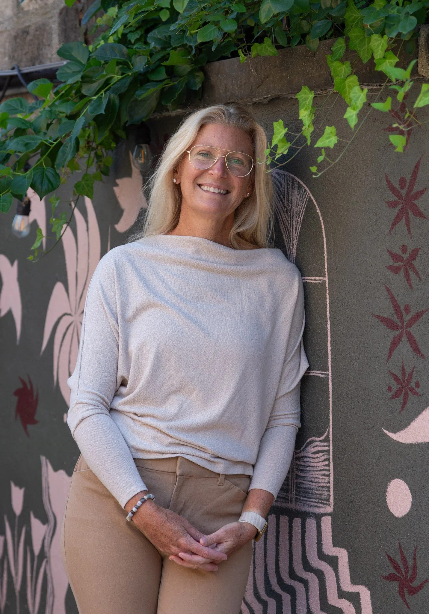 A smiling middle-aged woman with glasses and blonde hair, wearing a white long-sleeve top and beige pants, is standing in front of a chalkboard wall with colorful designs and ivy growing nearby.