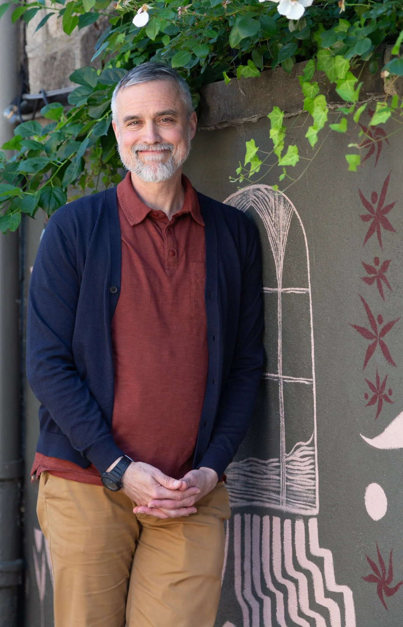 A smiling middle-aged man with gray hair and beard standing in front of a wall with chalk art of a window and flowers, wearing a red shirt, navy cardigan, khaki pants, and a wristwatch.