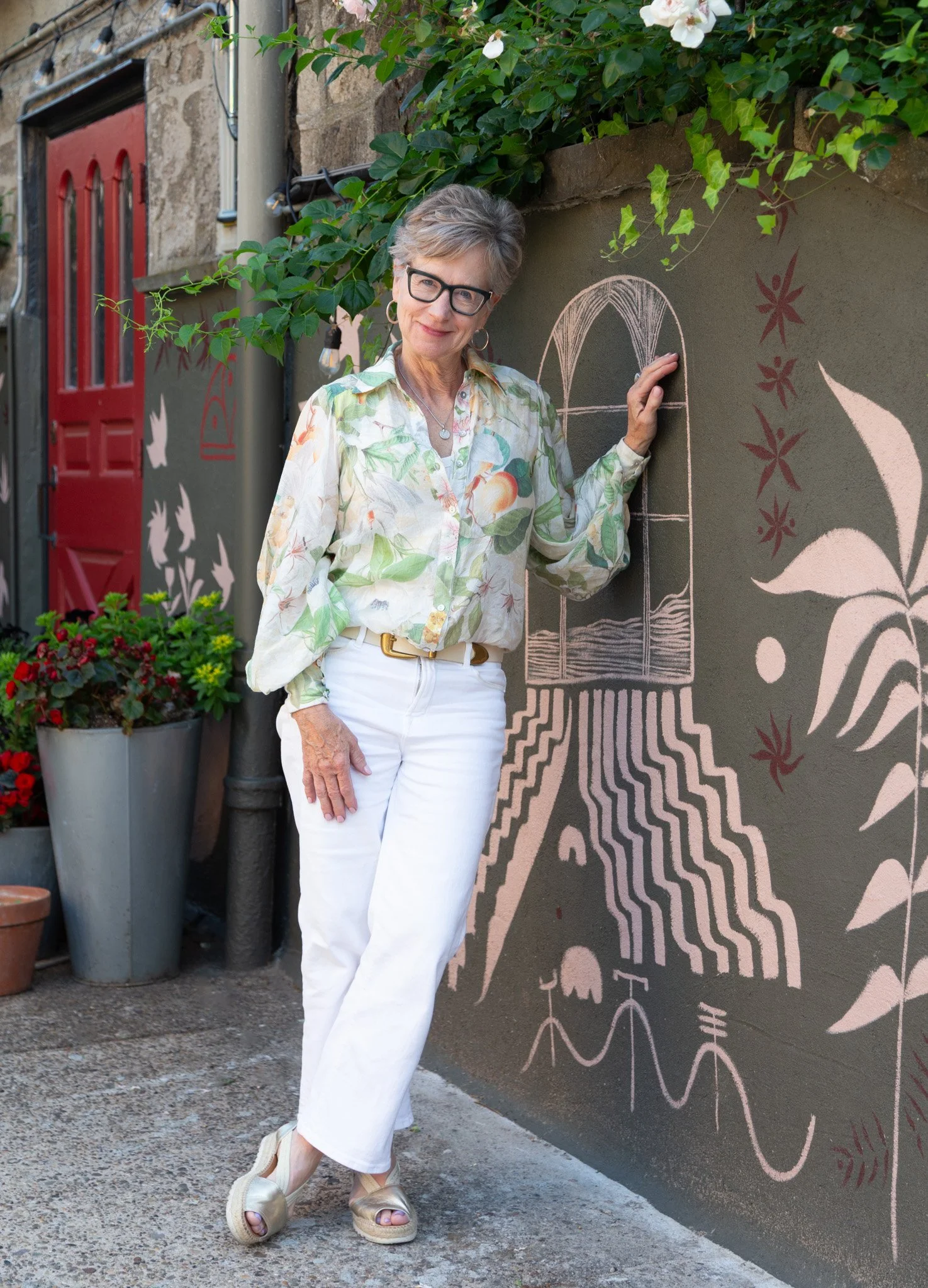 An elderly woman with short gray hair, glasses, and hoop earrings poses in front of a decorative wall with pink chalk art. She wears a light-colored, long-sleeved blouse with a fruit and leaf pattern, white pants, and tan wedge sandals. Green foliage and flowers are visible in the background.