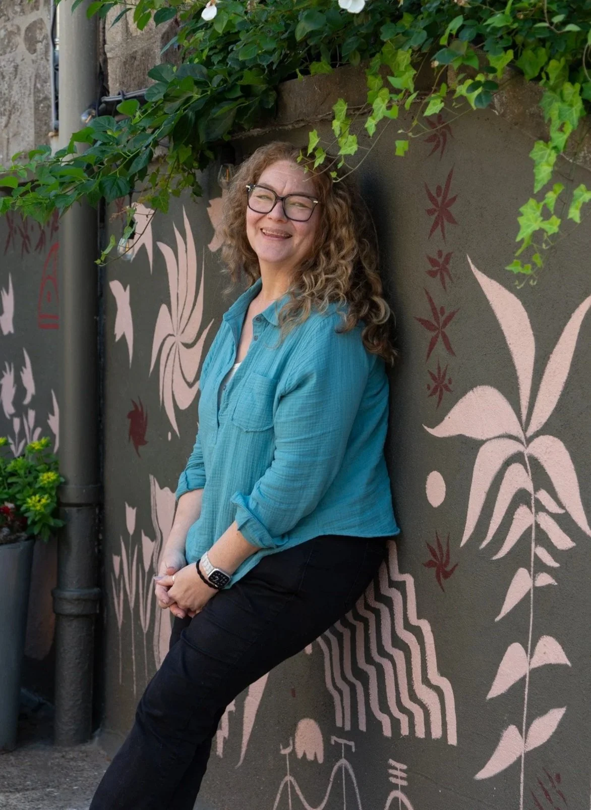 A woman with curly hair, glasses, and a blue shirt leaning against a wall with painted floral and geometric designs, smiling outdoors.