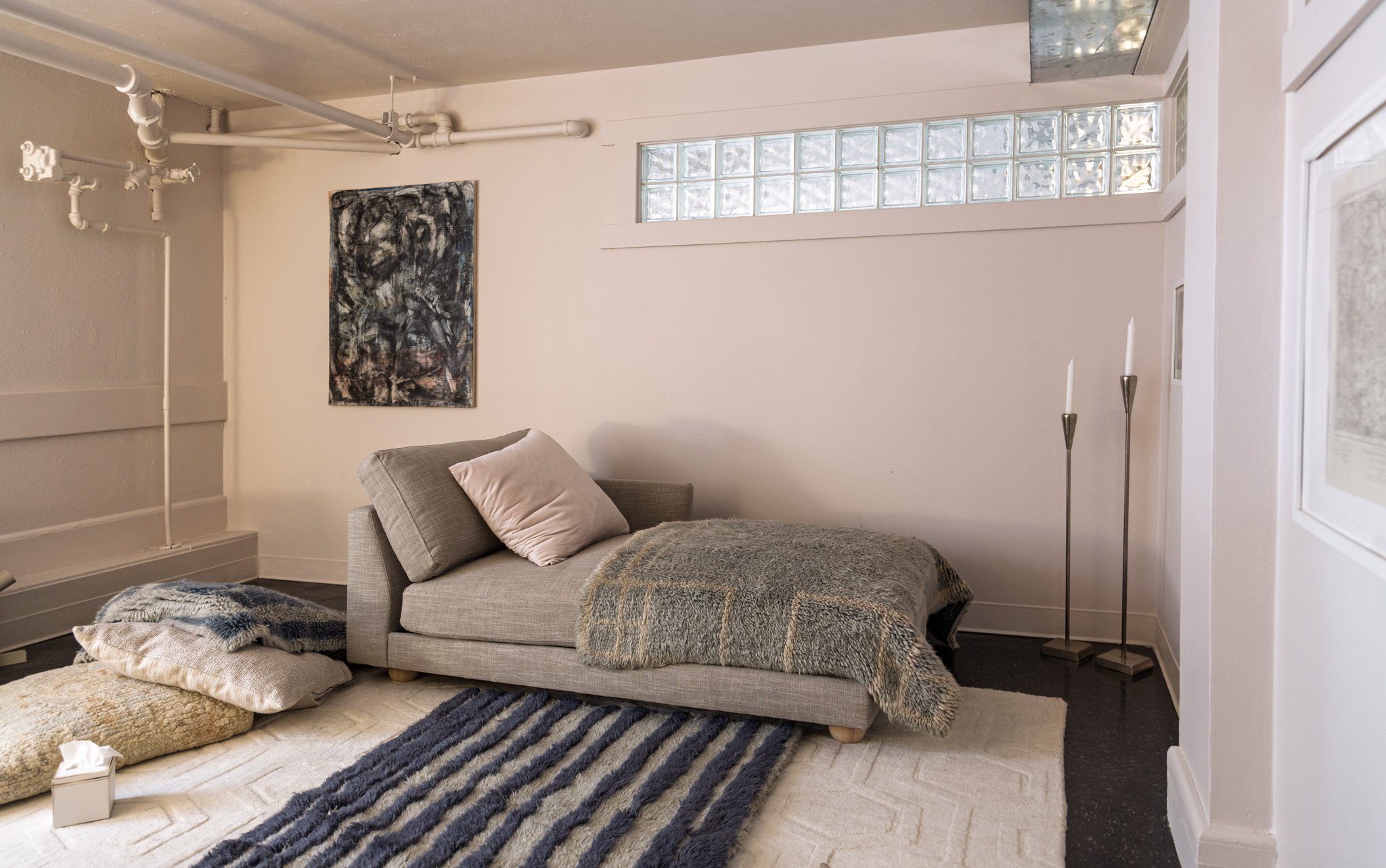 Living room with a gray couch, blankets on the floor, a wall painting, a glass block window, and tall candle holders.
