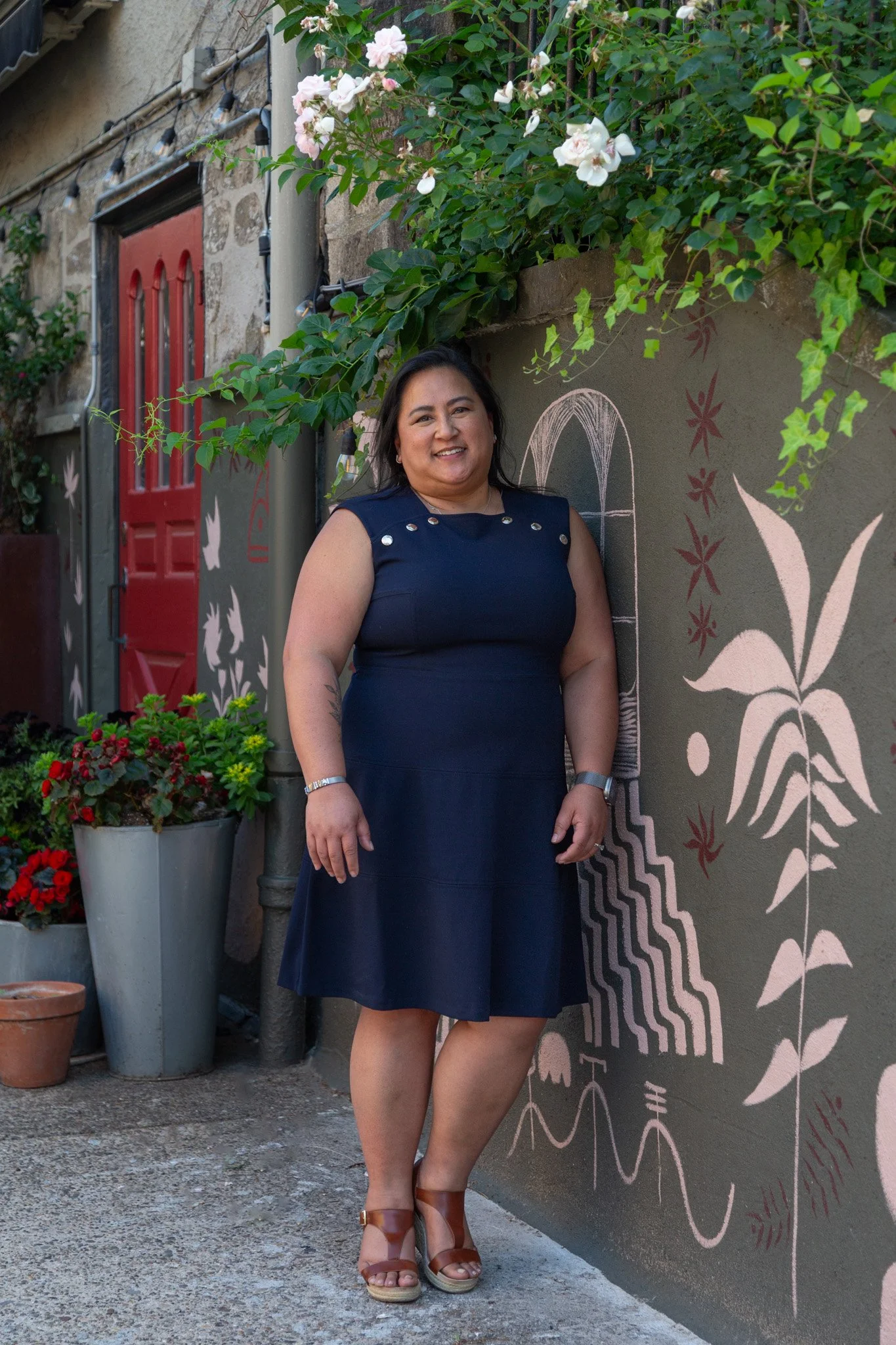A woman with dark hair in a navy blue dress, smiling, standing outdoors next to a mural on a wall with painted plants and abstract designs. She is wearing tan heels and a watch, with potted flowers nearby.