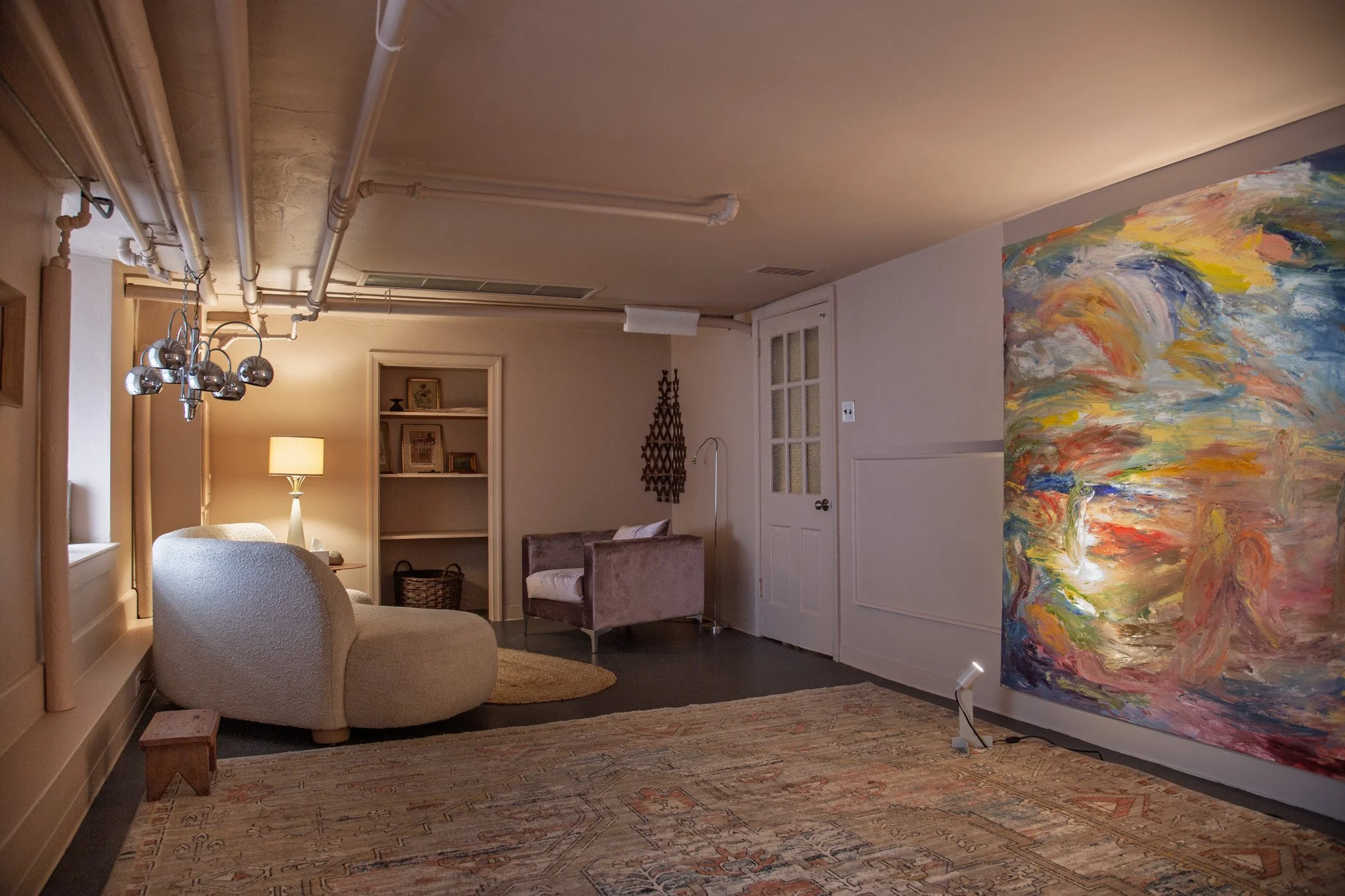 Living room with beige walls and contemporary furniture, including a rounded beige sofa, a basket, and an armchair. There is a large, colorful abstract painting on an easel. A patterned area rug covers the floor, and a table lamp and decorative wall hangings are visible.
