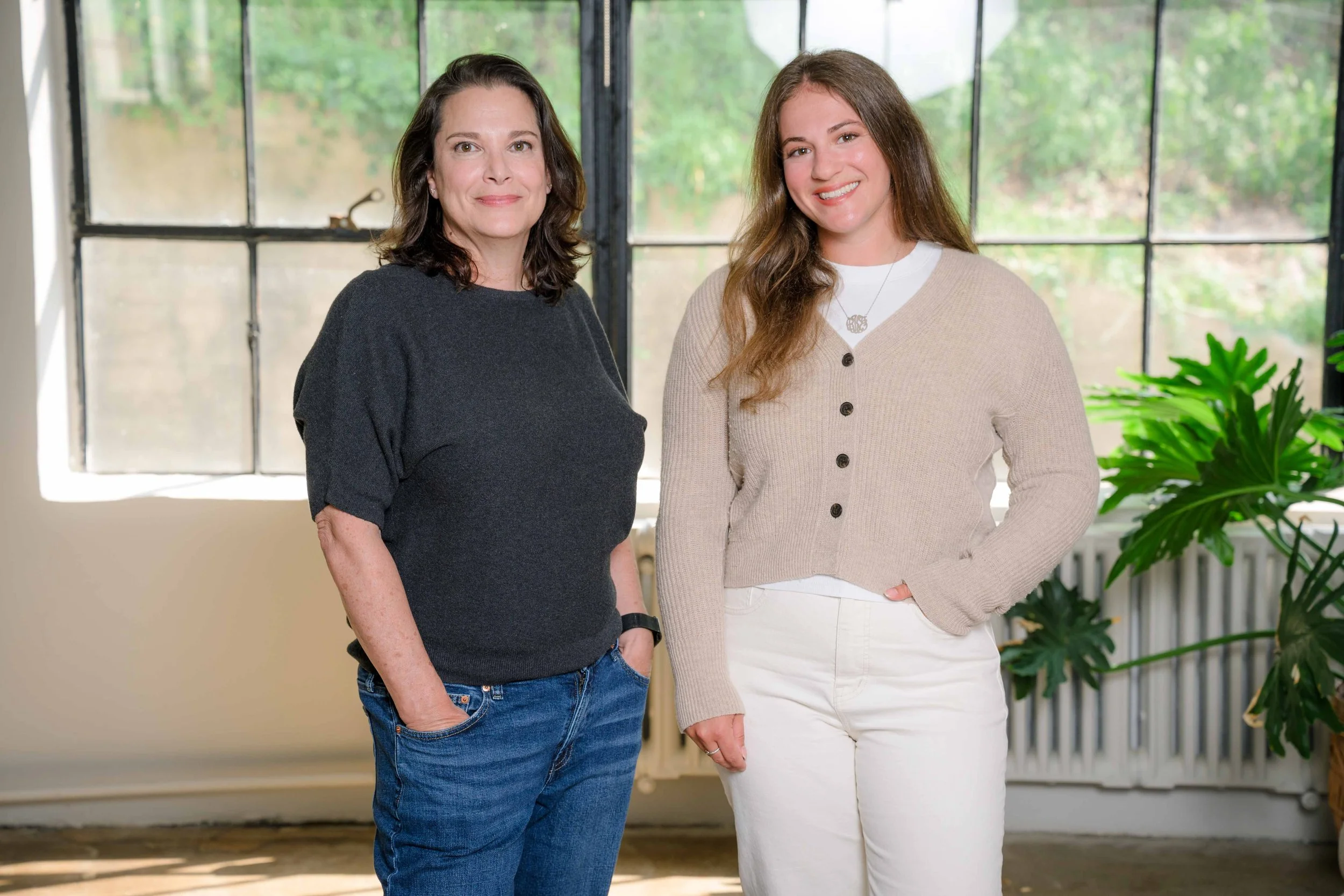 Two women standing inside near a window with greenery outside, smiling at the camera.
