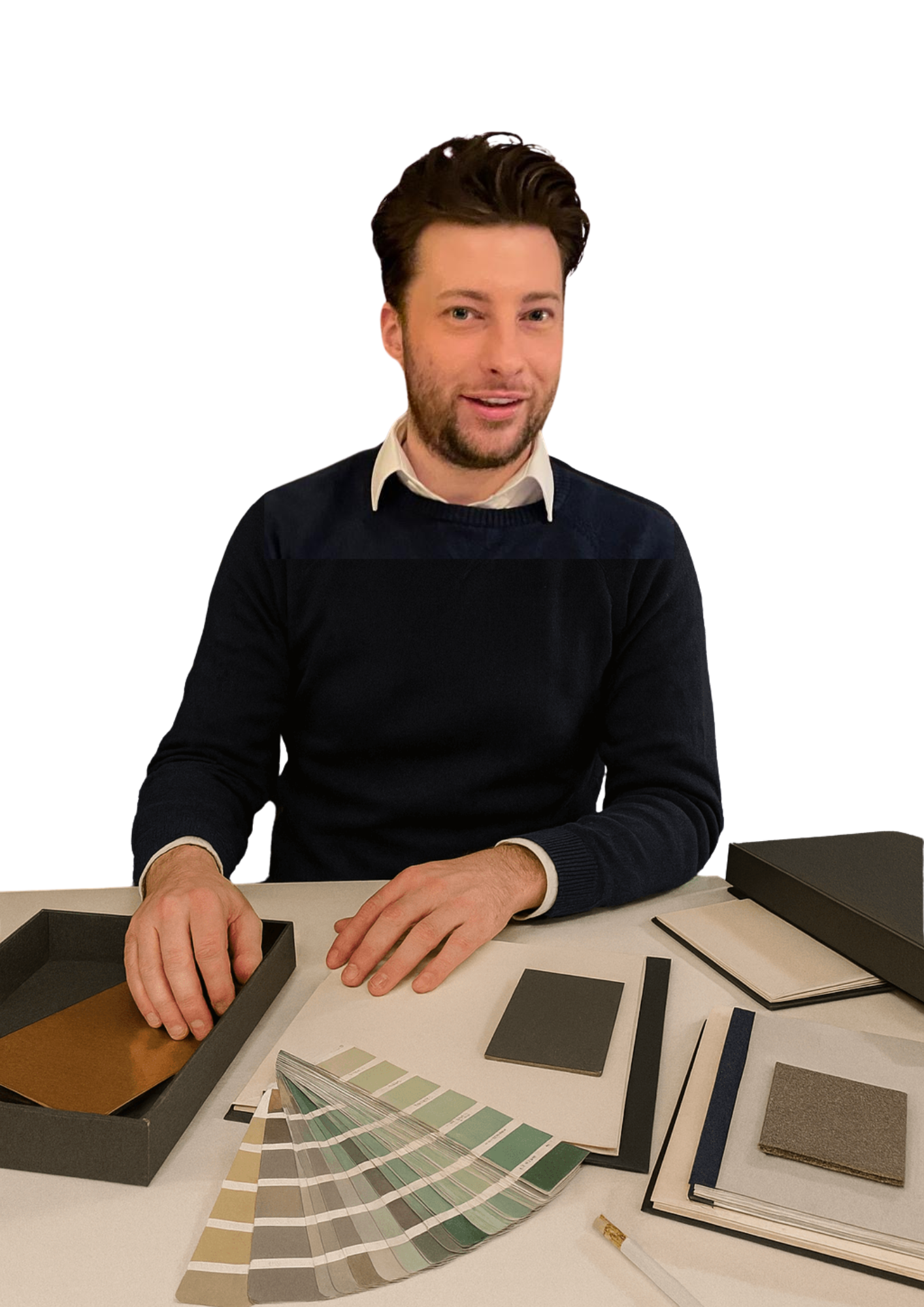 A man sitting at a desk with color swatches, fabric samples, notebooks, and a black box, smiling at the camera.