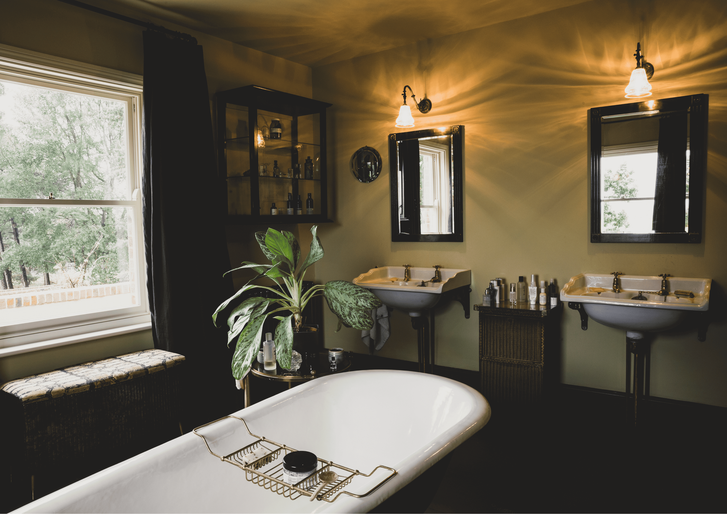 A vintage-style bathroom featuring a white bathtub, two wall-mounted sinks with brass fixtures, and two black-framed mirrors. The room has framed windows with black curtains, a small black cabinet with toiletries, a plant on a side table, and warm lighting from wall sconces, creating a cozy ambiance.