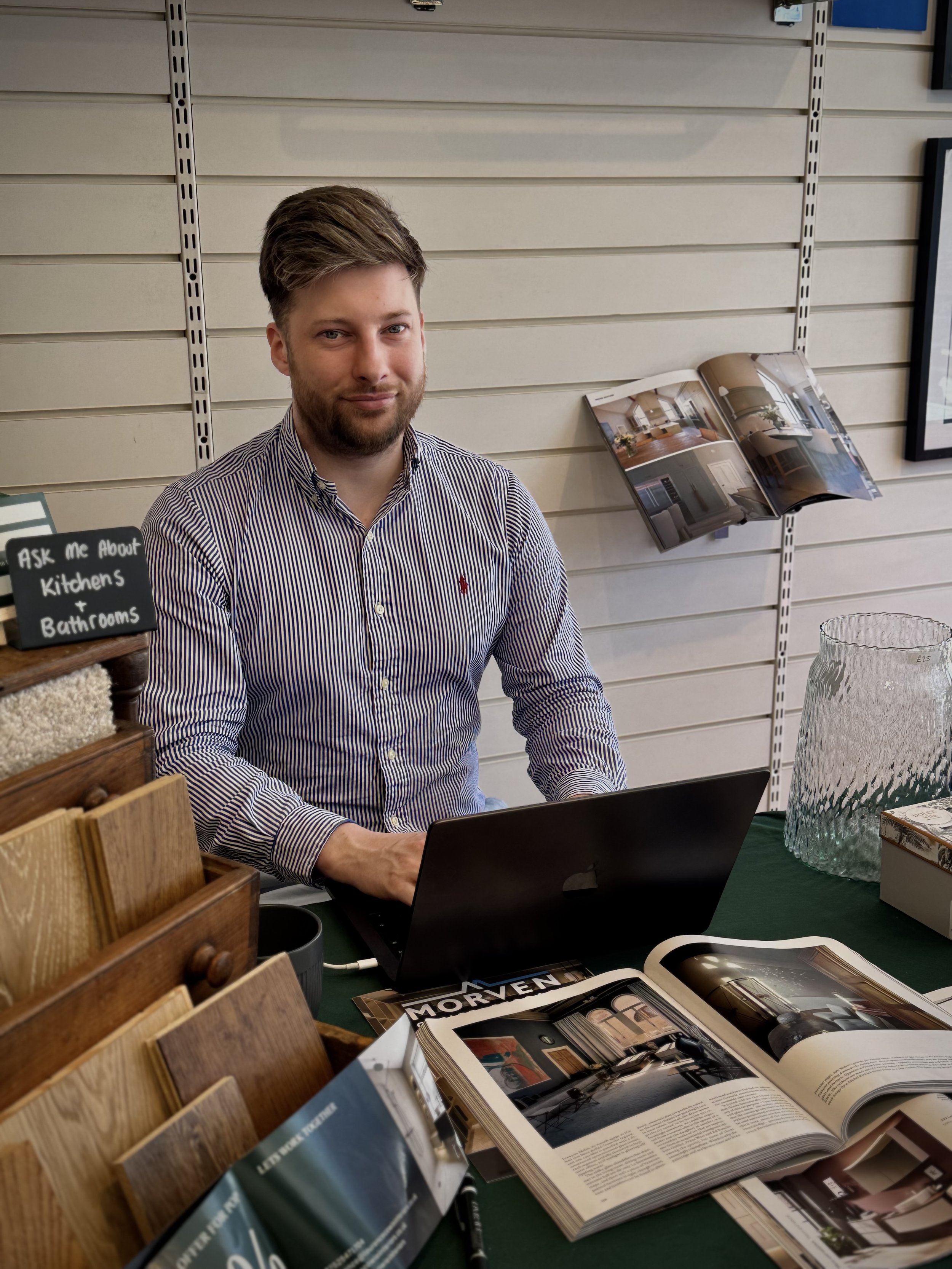 A man with a beard sitting at a table with a laptop, open magazines, and sample wood flooring samples displayed in front of him, in an interior design store.