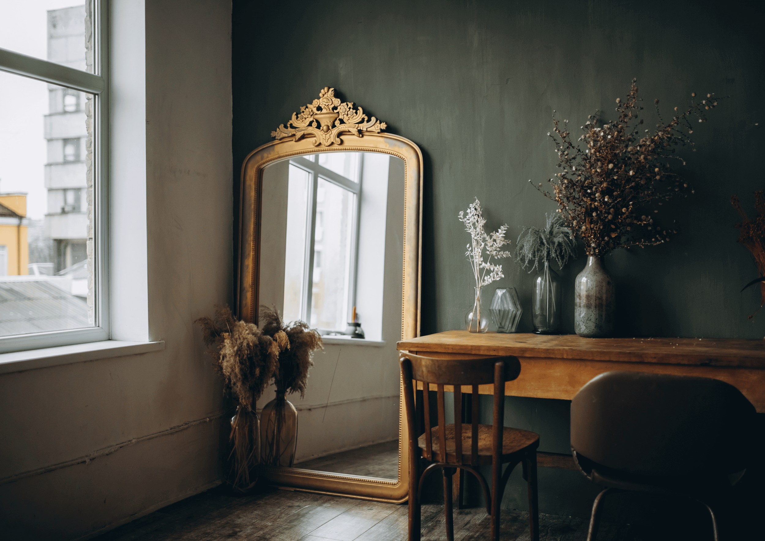 Interior of a room with a tall ornate mirror, a wooden table, and vases with dried flowers, near a window with a cityscape view.