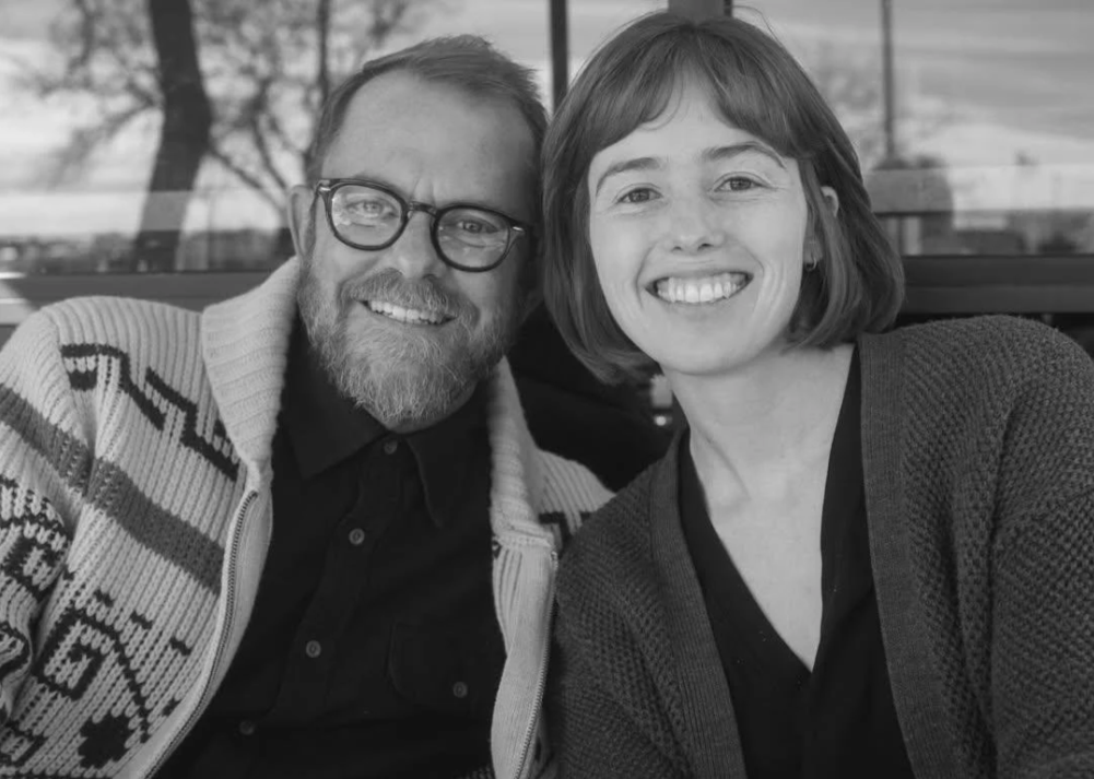A black-and-white photo of a man and woman sitting close together, smiling at the camera, outdoors near a window with trees visible in the background.