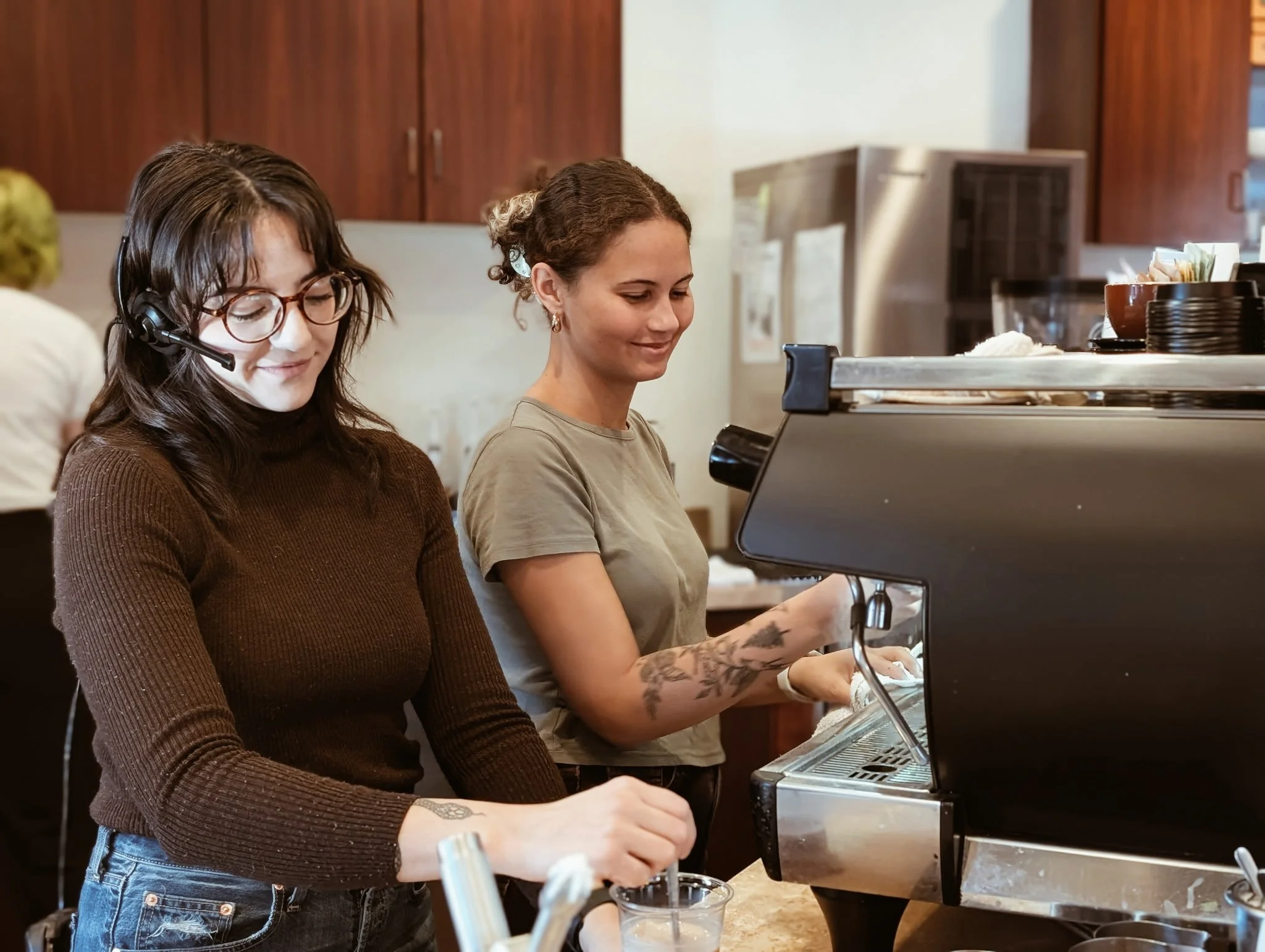 Two women making coffee at a coffee shop, one using an espresso machine, with a wood-paneled wall in the background.