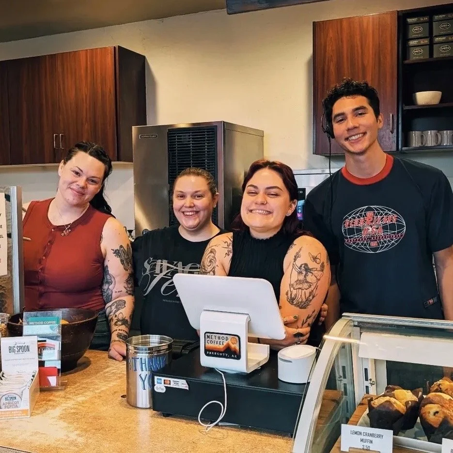 Four smiling staff members behind the counter at a coffee shop, with a display case of muffins in front.