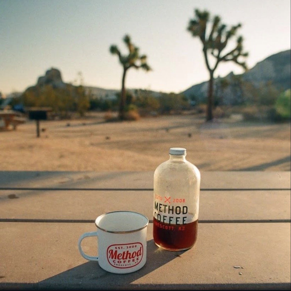 A coffee mug and a bottle of Method Coffee on a table in a desert landscape with Joshua trees and hills in the background, during sunset.