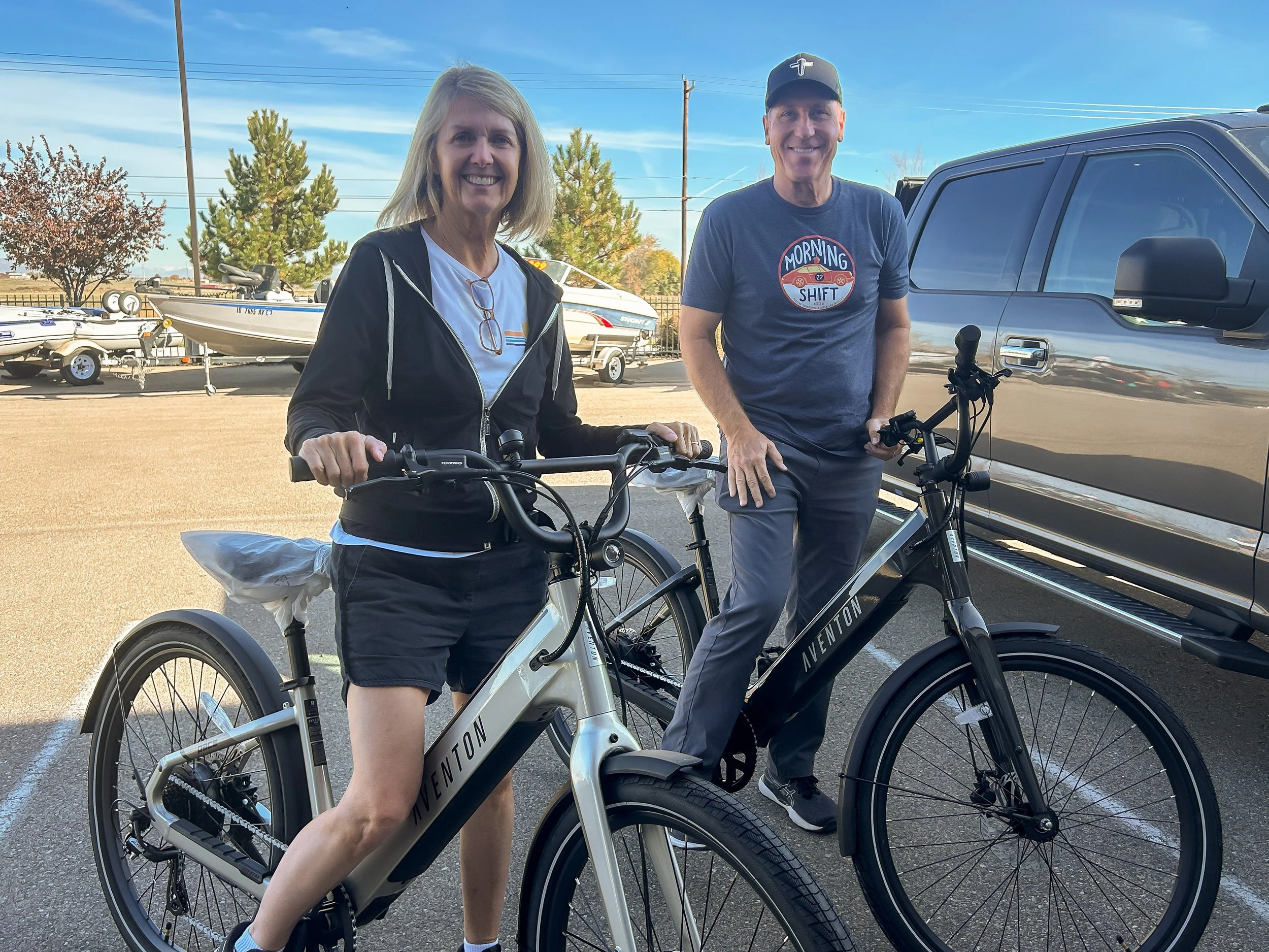 A woman and a man standing with electric bicycles, smiling outdoors with boats and trees in the background.
