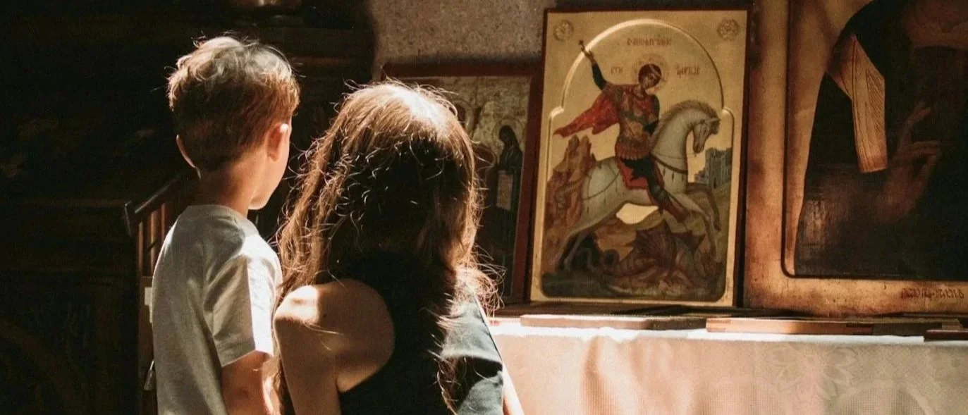 Two children, a boy and a girl, observing religious Orthodox Christian icons displayed on a wall.