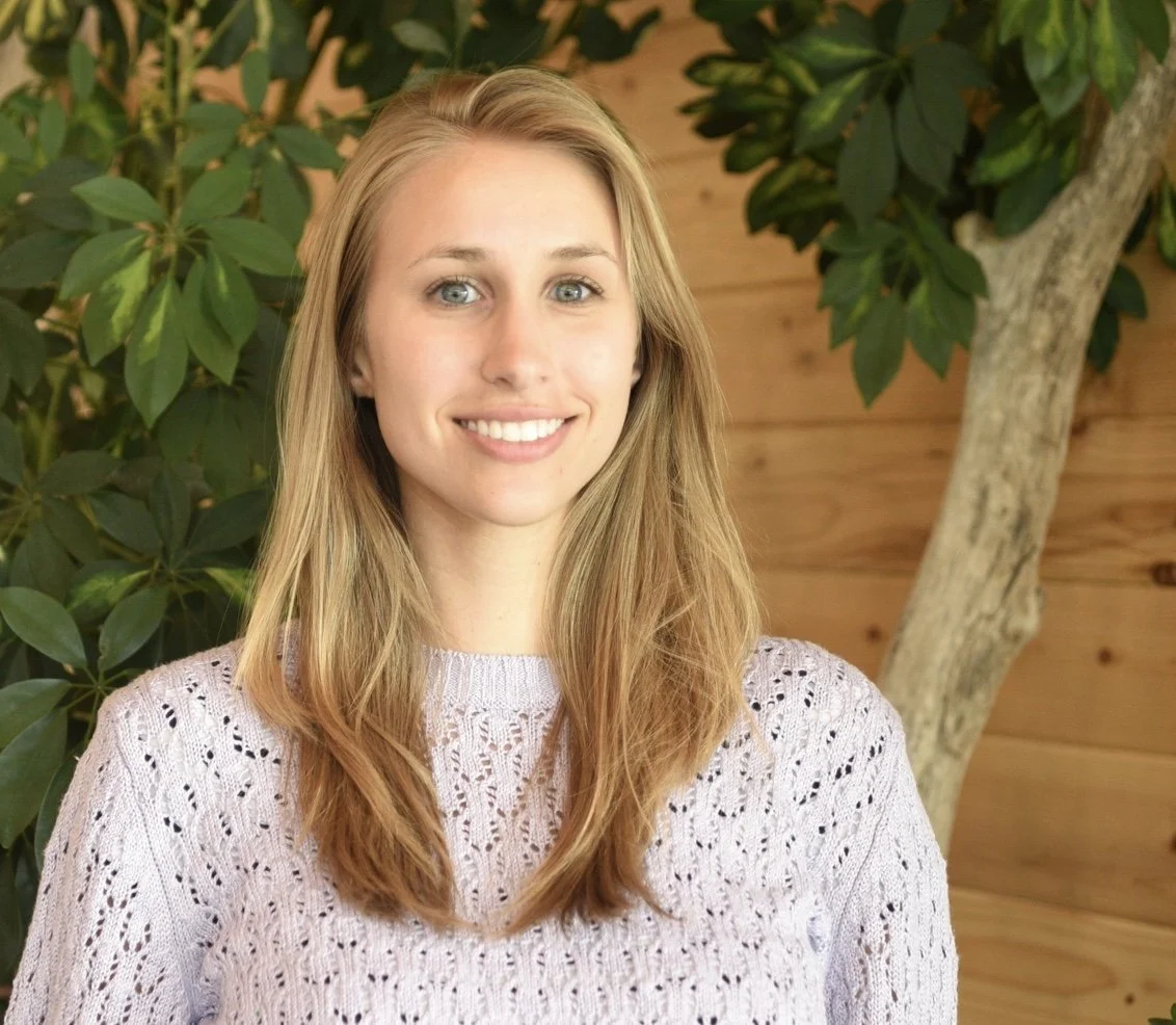 Woman with long blonde hair wearing an purple knit sweater, looking at the camera against a foliage and wood paneling background.