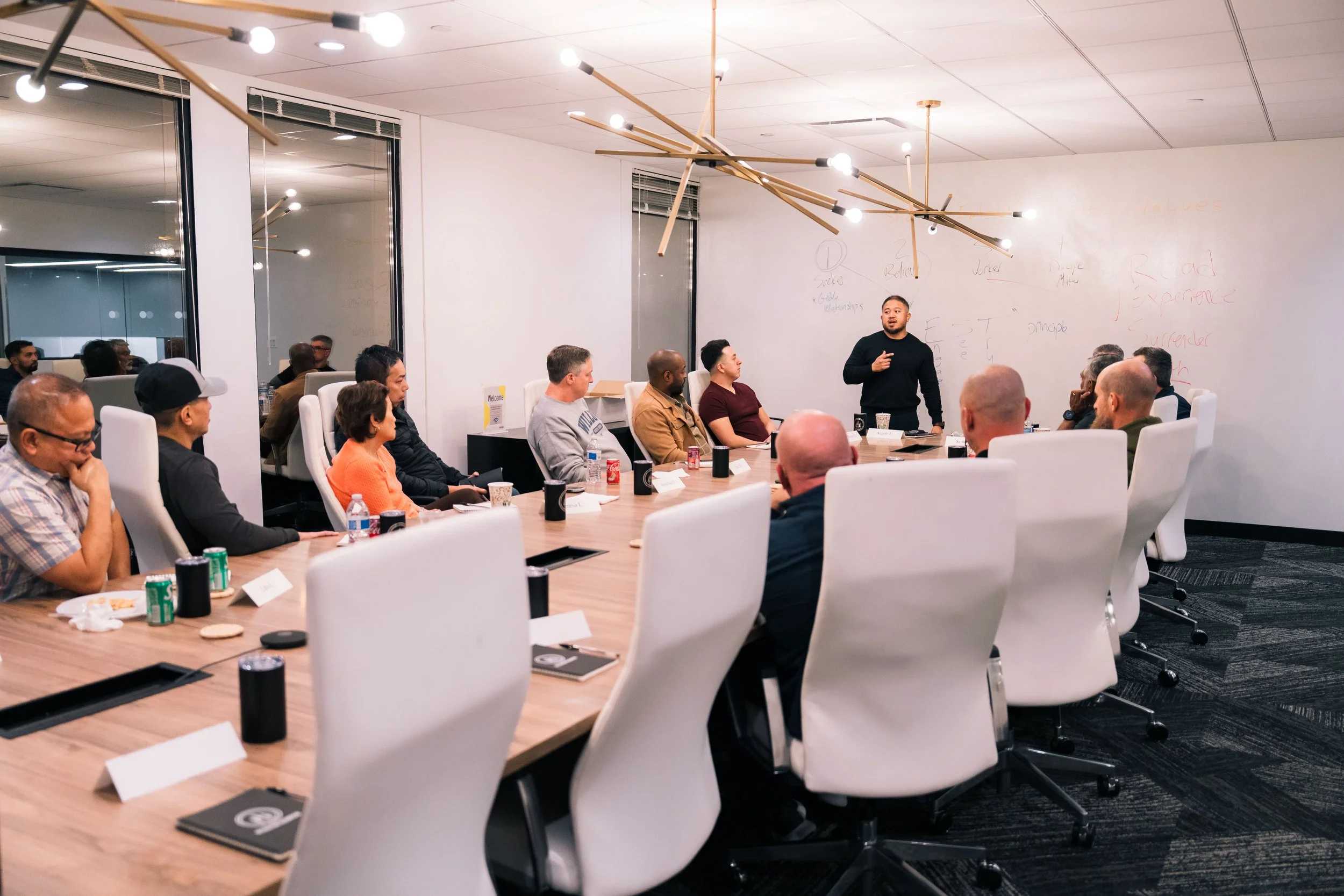 A man giving a presentation in a conference room with a large whiteboard and an audience seated around a conference table.