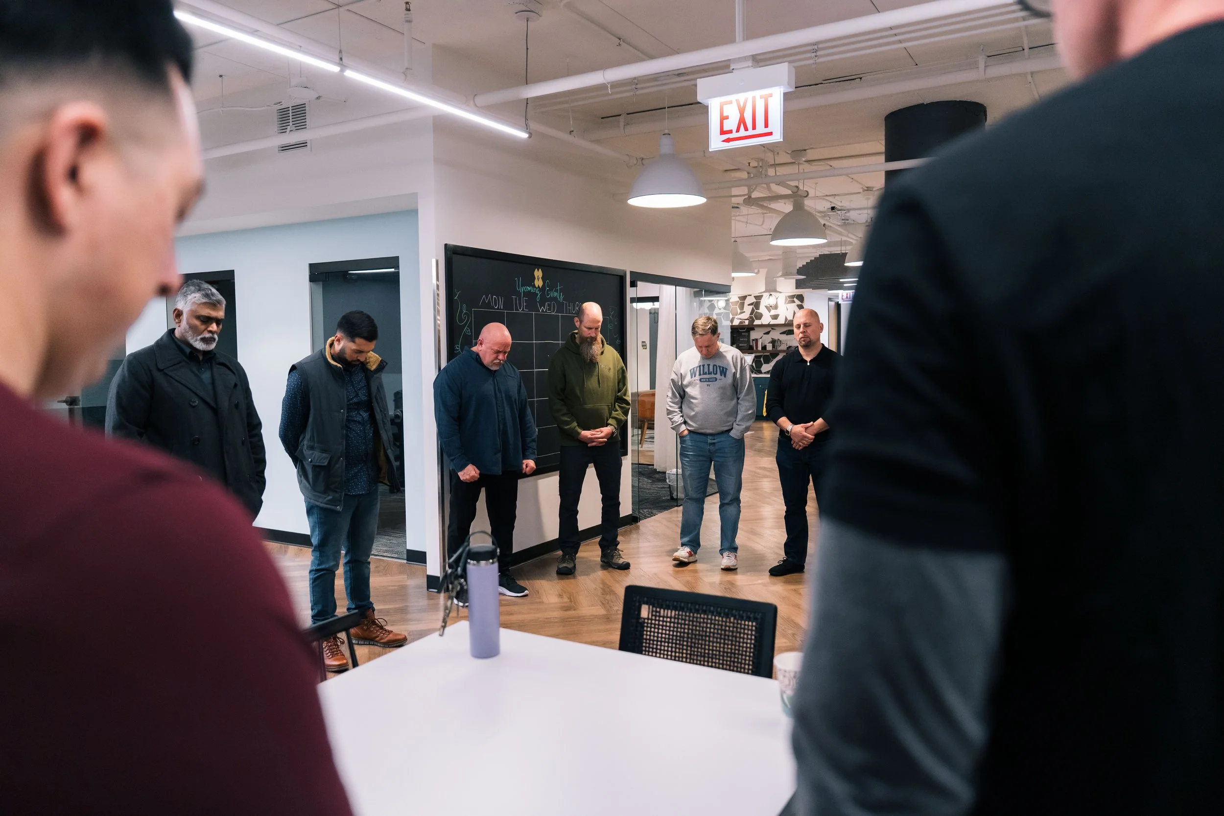 A group of seven men standing with their heads bowed and eyes closed in a circle inside an office or meeting space.