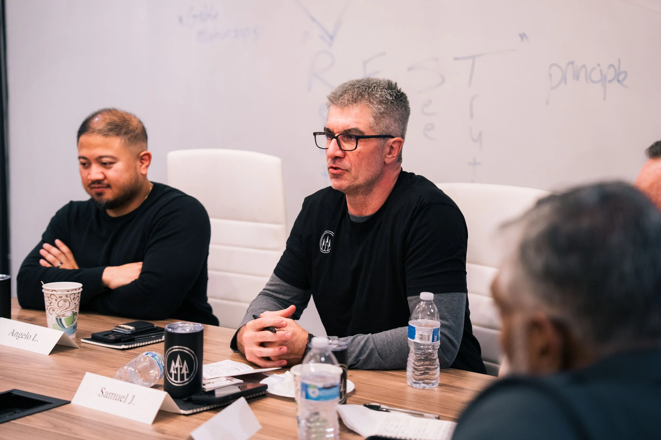 People sitting at a conference table during a meeting, with water bottles, notebooks, and drinks in front of them, and a whiteboard in the background.