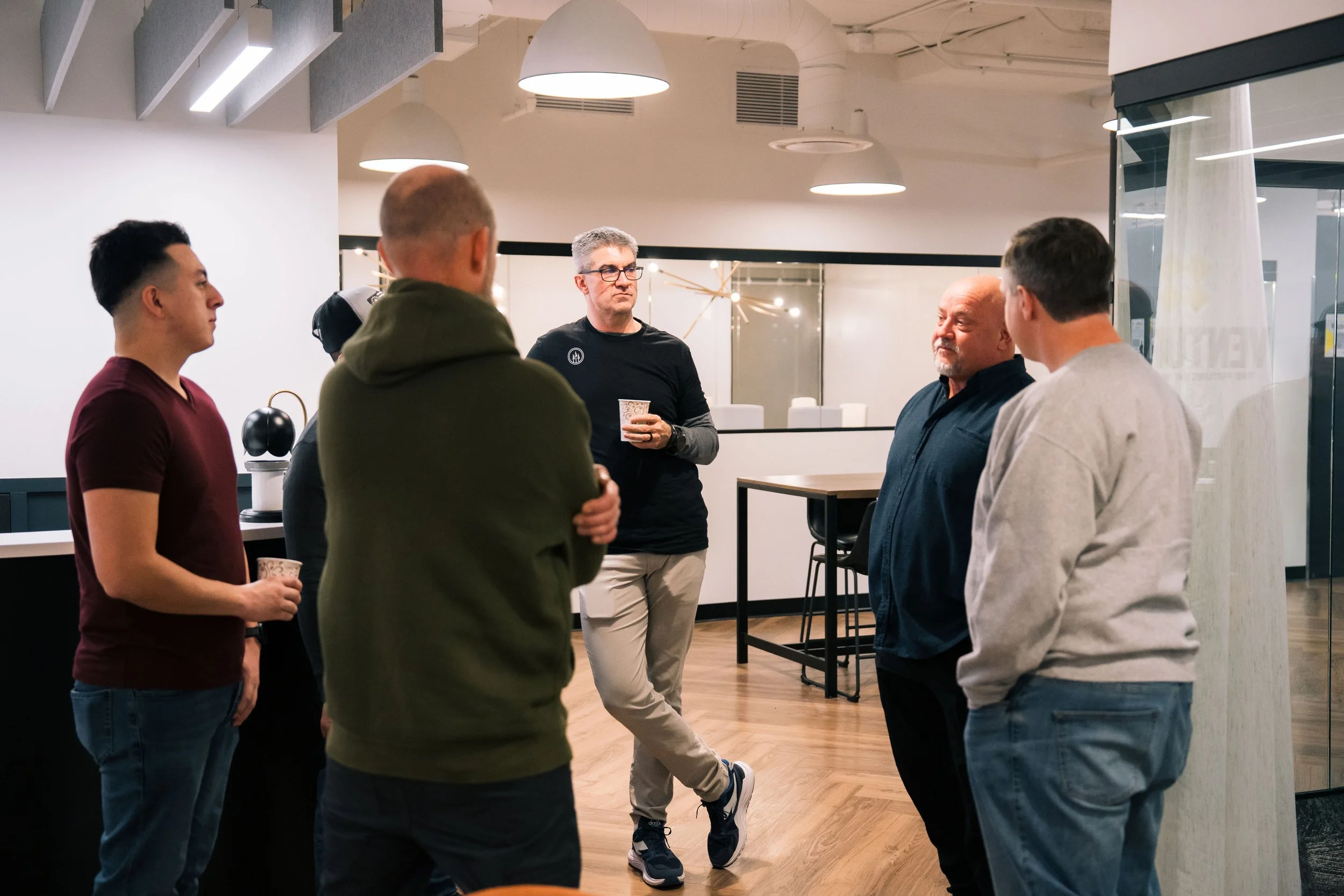 Group of five men in casual attire having a conversation in an indoor modern office space with wooden floor and bright lighting.