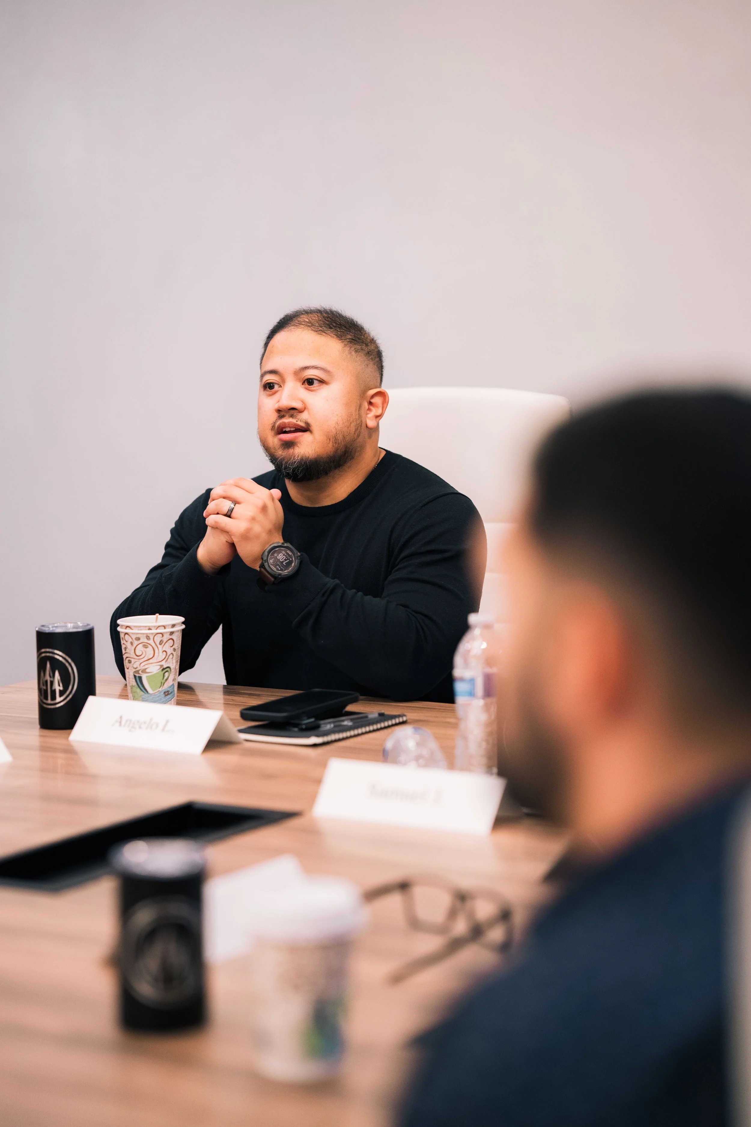 Man sitting at a conference table, speaking and with hands clasped, wearing a black long-sleeve shirt, with notebooks, cups, and a water bottle in front, and a name tag that reads Angela L.