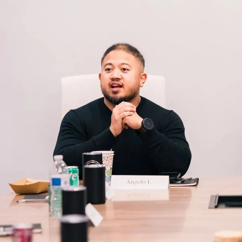 Man with short hair and beard wearing a black shirt, sitting at a conference table with various items including cups, cans, and a phone. He appears to be speaking or listening during a meeting.