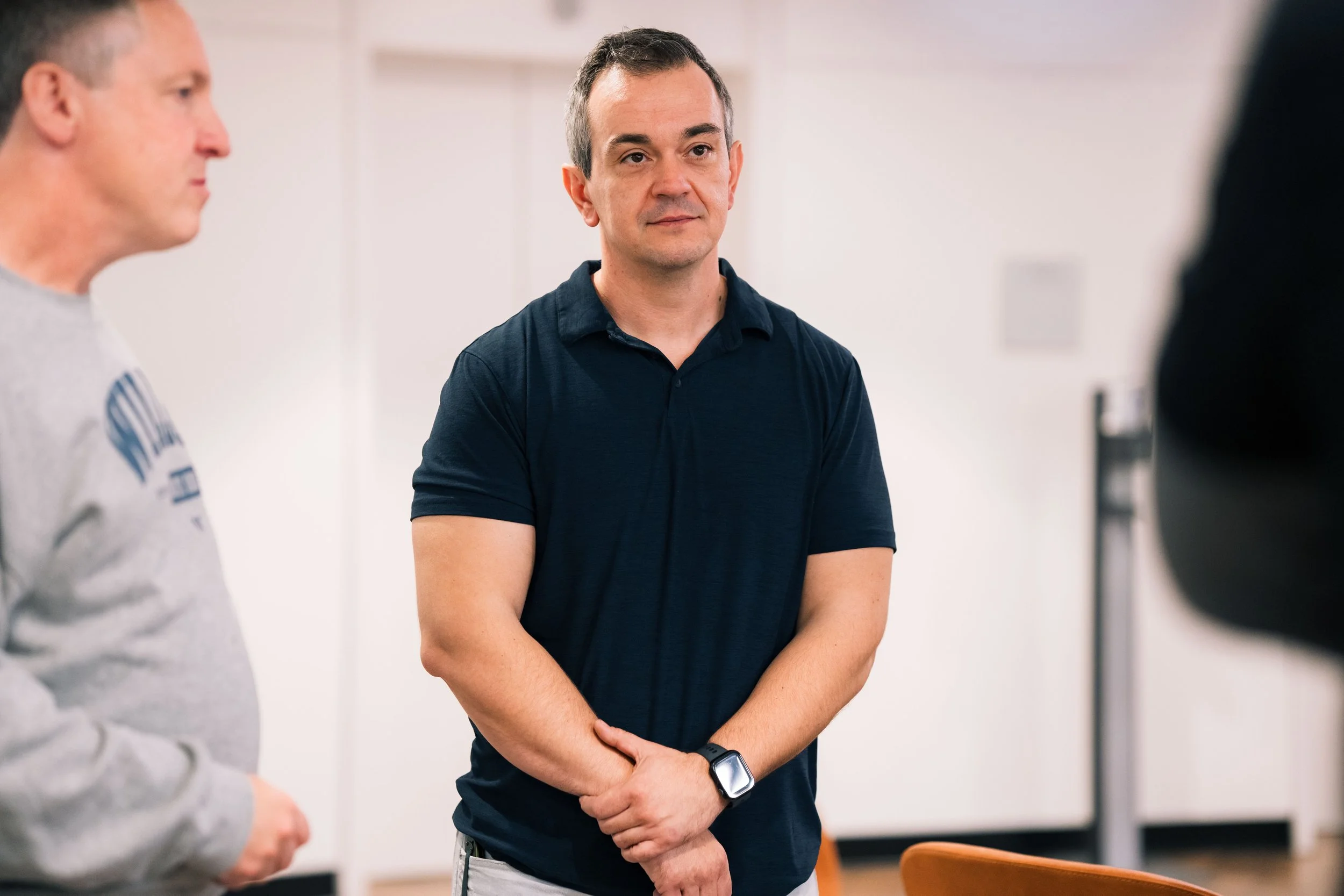 A man in a black polo shirt standing with arms crossed and looking contemplative, with two other men partially visible on either side, indoors in a well-lit room.