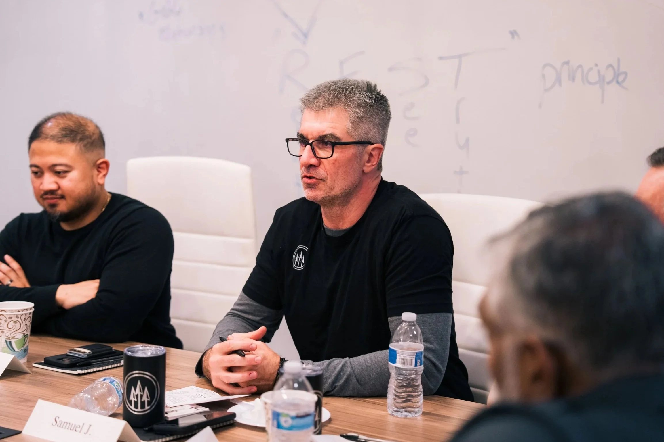 A group of men sitting around a conference table, engaged in discussion, with whiteboard in the background.