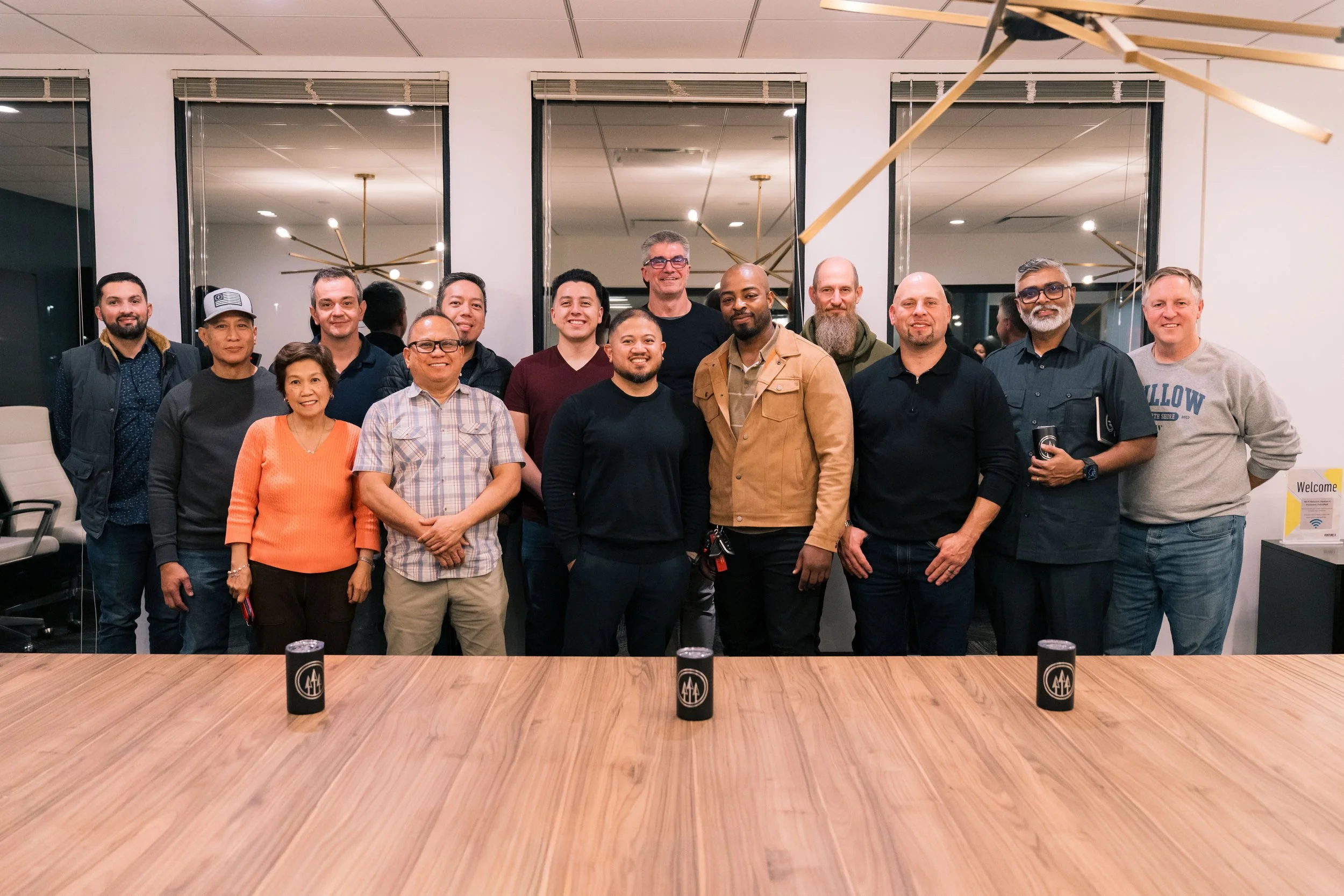 Group of 14 people standing together in an indoor setting, smiling for a photo, behind a wooden table with three black cups on it.